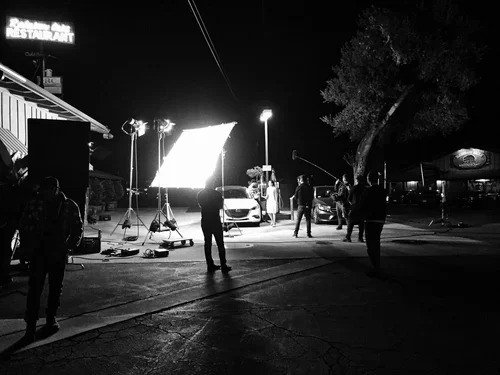 Film crew working on a night shoot with lighting equipment, a parked car, and crew members in an outdoor parking lot or gas station.