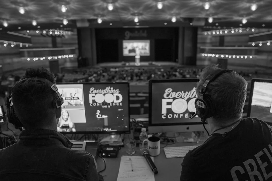A video production team at a food conference, sitting at a table with monitors displaying the conference branding and presentation, with an audience in the auditorium.