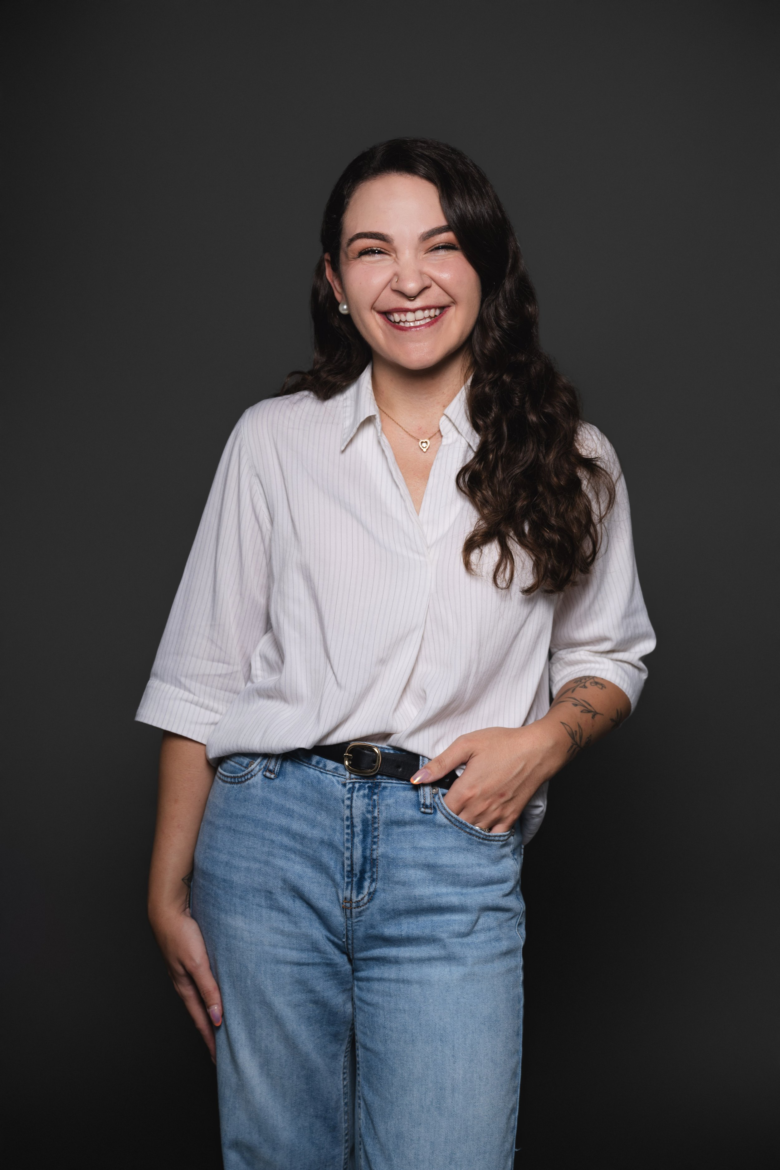 A woman with long, wavy dark hair, wearing a white striped button-up shirt, blue jeans, and jewelry, smiling against a dark background.