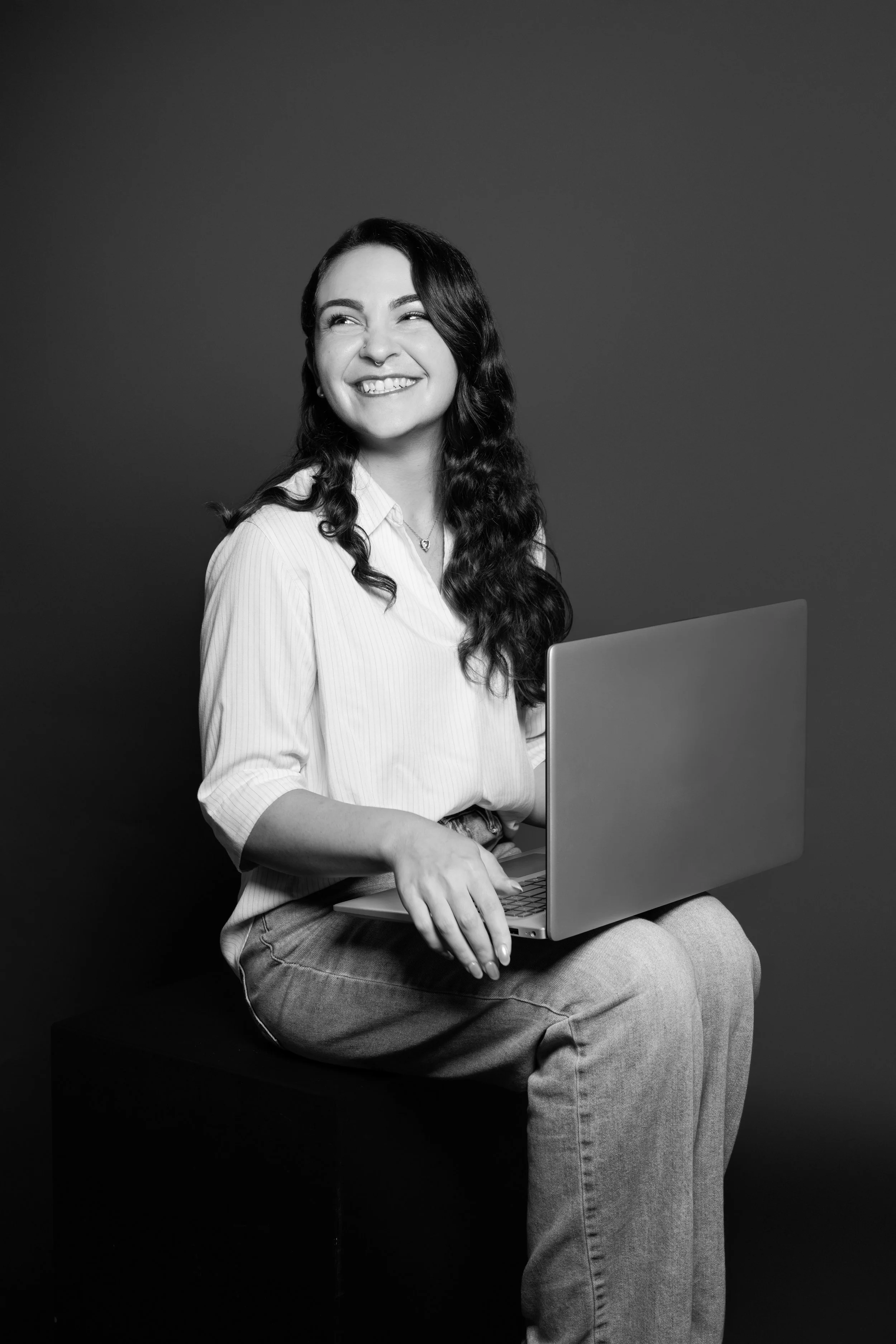 A young woman with wavy dark hair, smiling and looking to the side, sitting on a stool with a laptop on her lap, against a dark background.