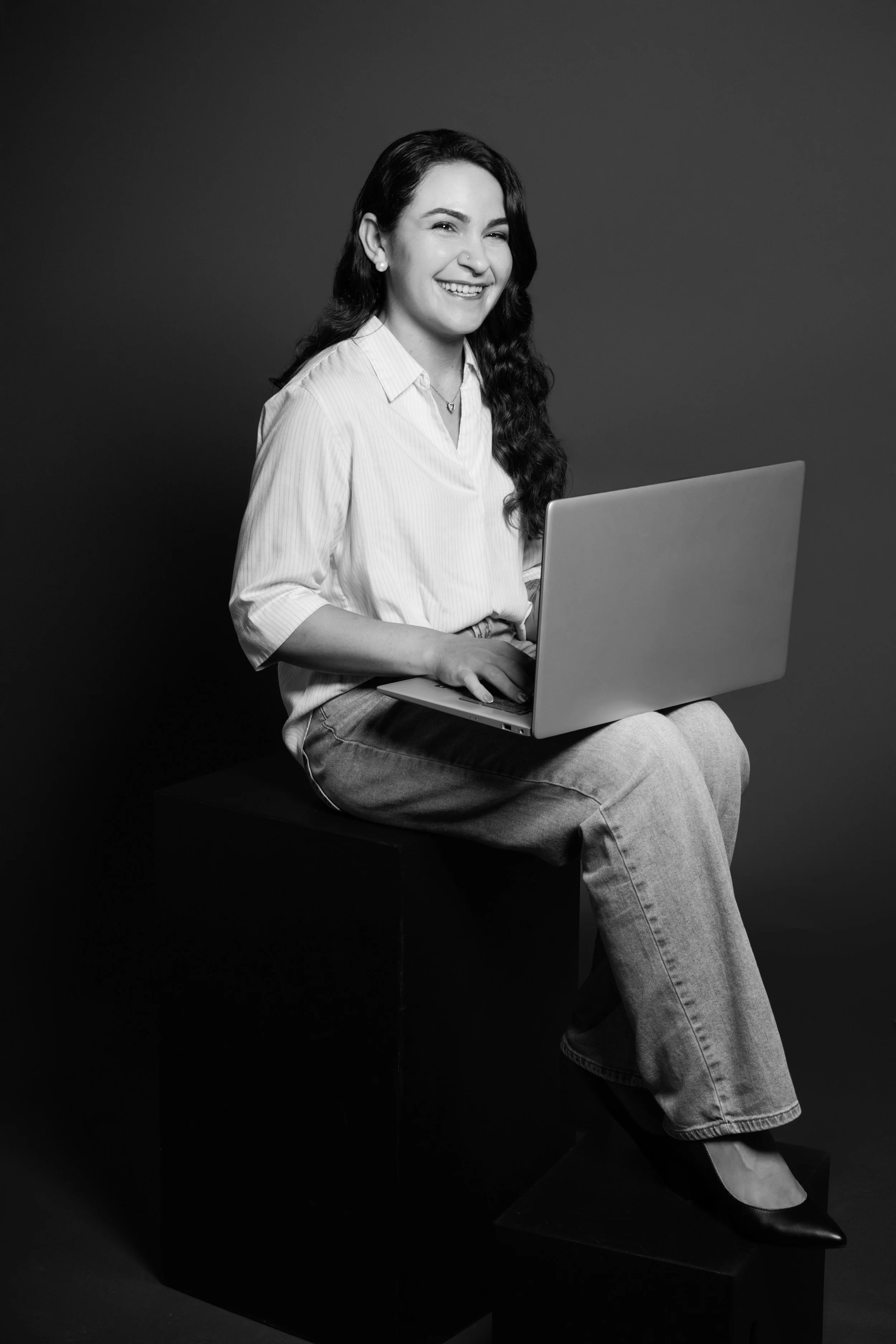 Black and white photo of a smiling woman with long curly hair, wearing a white blouse, sitting on a black cube, working on a laptop against a dark background.