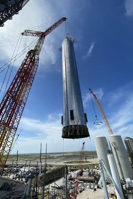 A rocket is being lifted by cranes at a launch pad, preparing for a launch against a blue sky with some clouds.