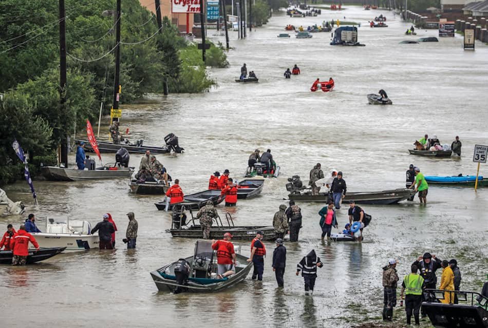 Numerous boats and rescue personnel navigating a flooded street with submerged vehicles and water reaching the buildings' entrances.