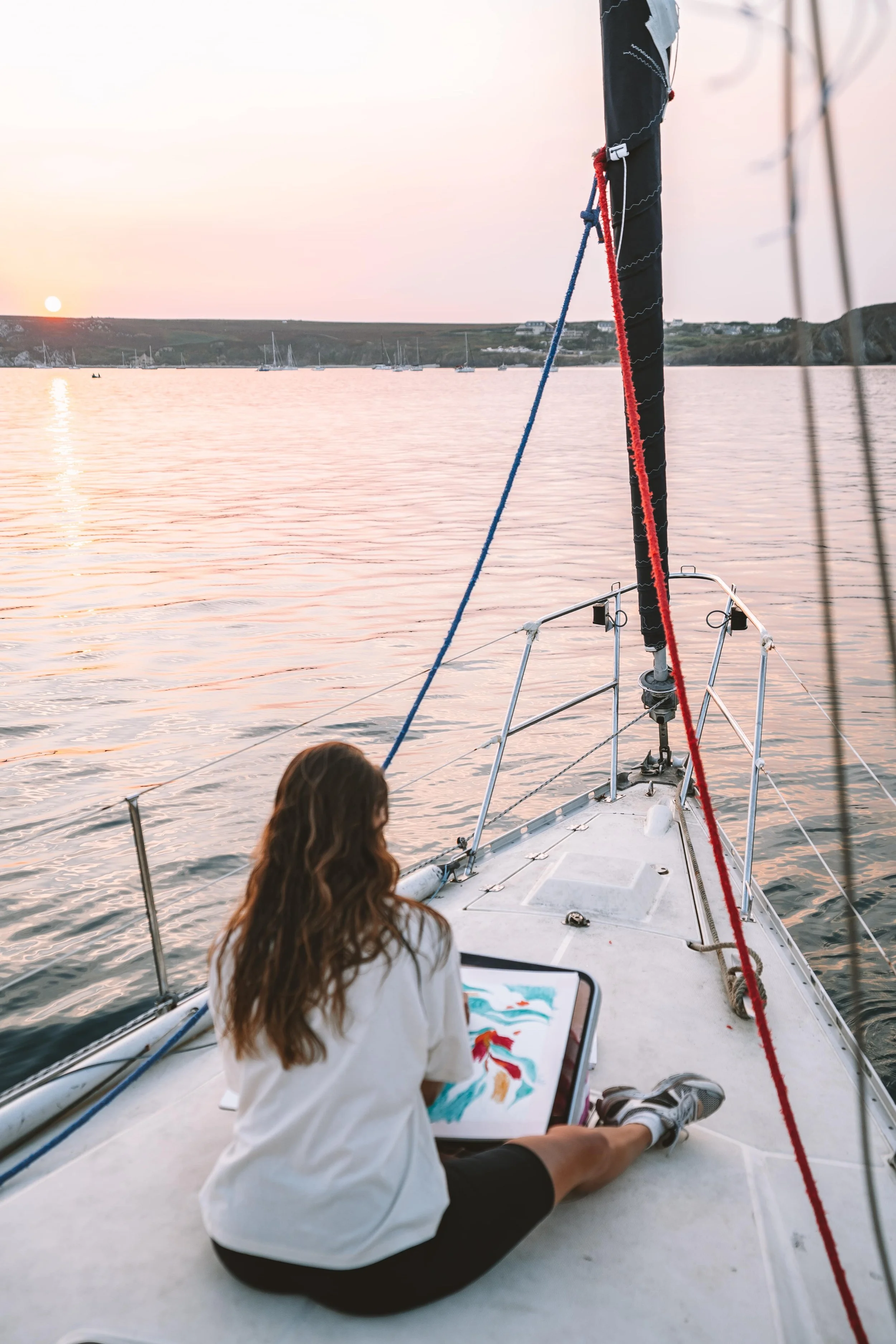 Une femme assise sur le pont d'un bateau au coucher du soleil, en train de peindre sur un carnet de croquis, avec une vue sur la mer et plusieurs voiliers en arrière-plan.