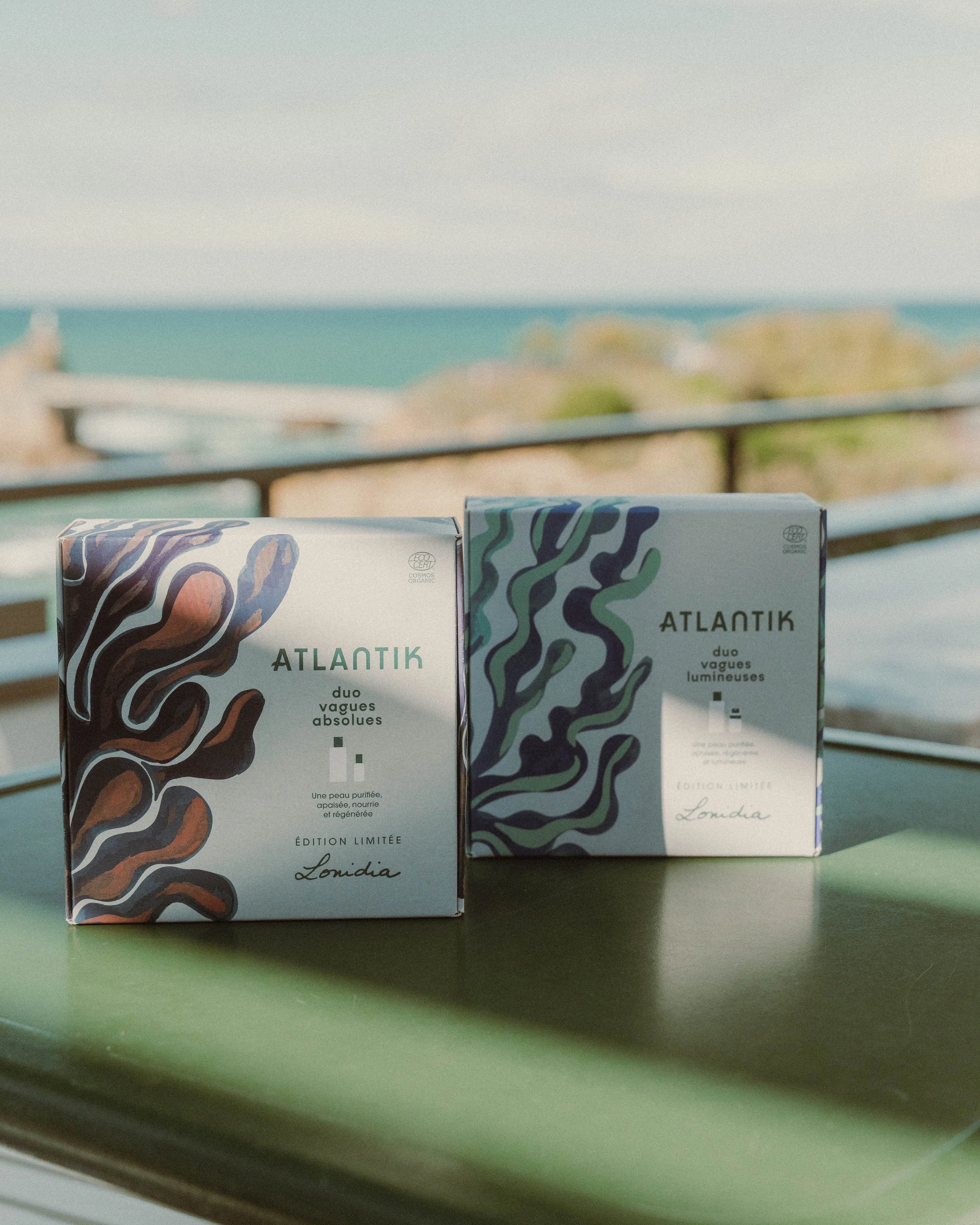 Deux boîtes de produits cosmétiques Atlantik Care avec des motifs colorés, posées sur une table devant un fond flou, probablement une scène en bord de mer ou en terrasse.