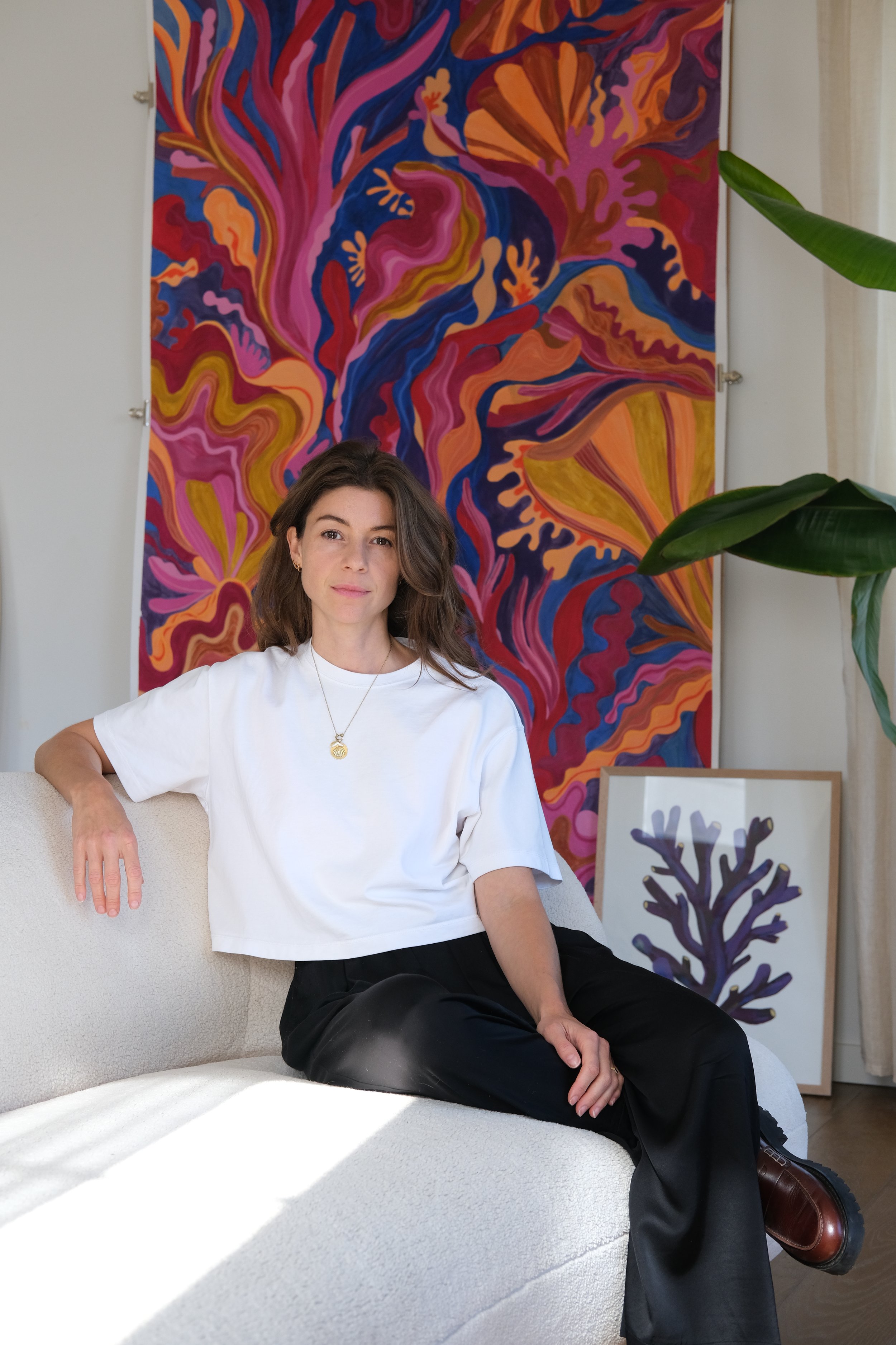 Une femme assise sur un canapé blanc dans un intérieur avec des œuvres d'art colorées en arrière-plan, portant un chemisier blanc, un collier en or avec un symbole et des pantalons noirs.