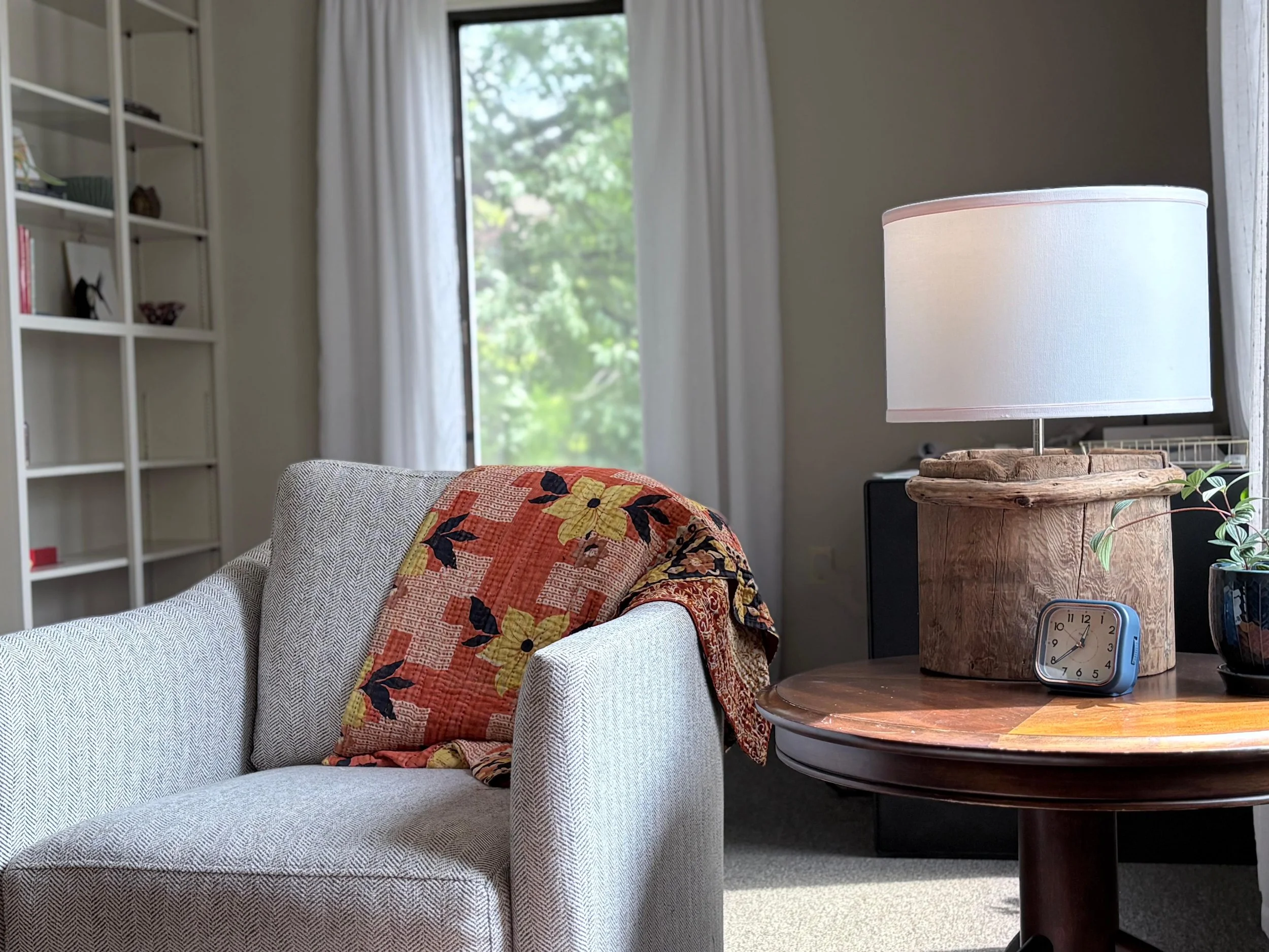 A cozy living room with a white sofa draped with a colorful quilt, a side table with a clock and plants, a lamp, a bookshelf, and a window with white curtains showing green foliage outside.