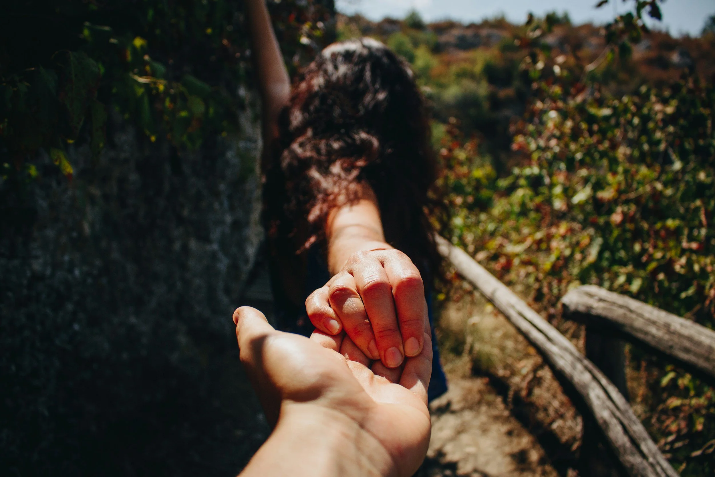 A person with long curly dark hair reaching out their hand, holding the hand of another person, outdoors with greenery and a fallen log in the background.