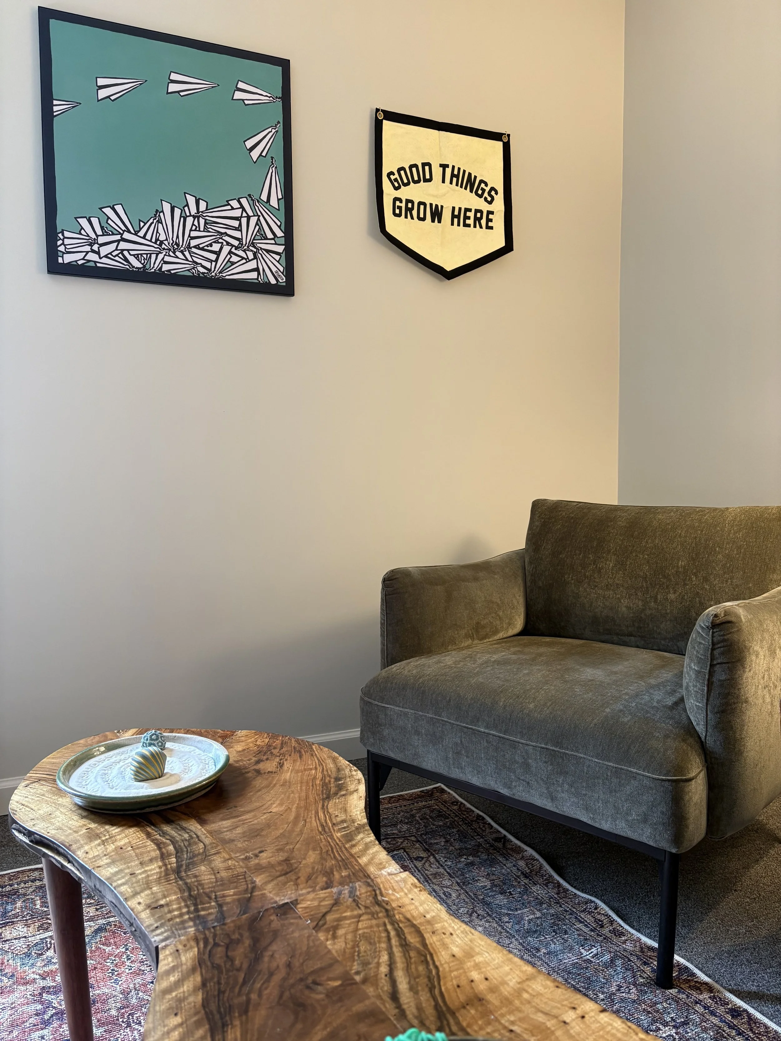 A living room corner with a dark grey upholstered sofa, a wooden coffee table with a bowl of decorative eggs, a patterned rug, and wall art including a painting of paper airplanes and a wall hanging that reads "Good Things Grow Here".