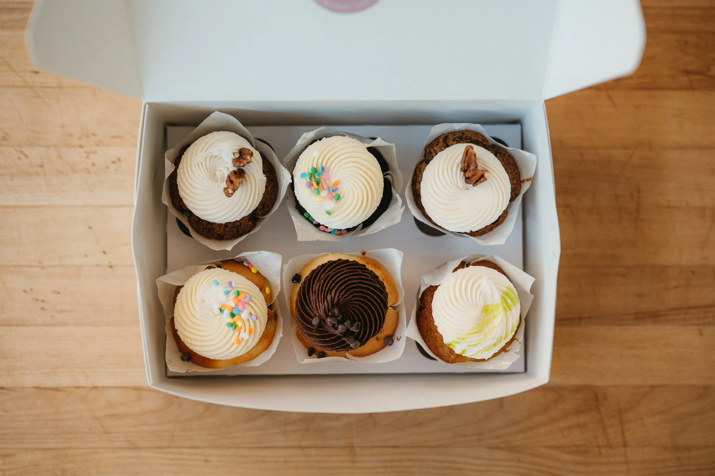 A box of assorted cupcakes topped with frosting, sprinkles, and nuts, placed on a wooden surface.