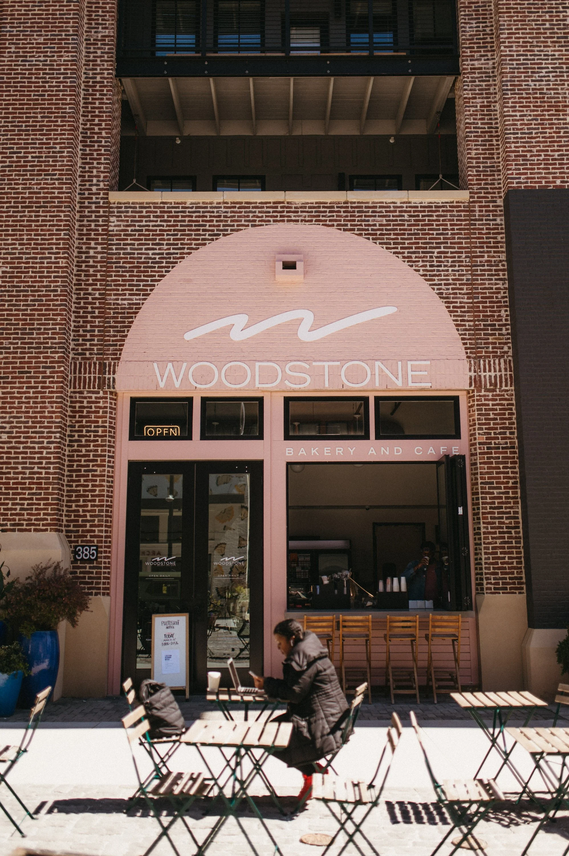 The image shows the storefront of Woodstone Bakery and Café. People are sitting outside at tables, with a woman working on her laptop. The building has a red brick exterior and pink accents around the entrance, with a neon