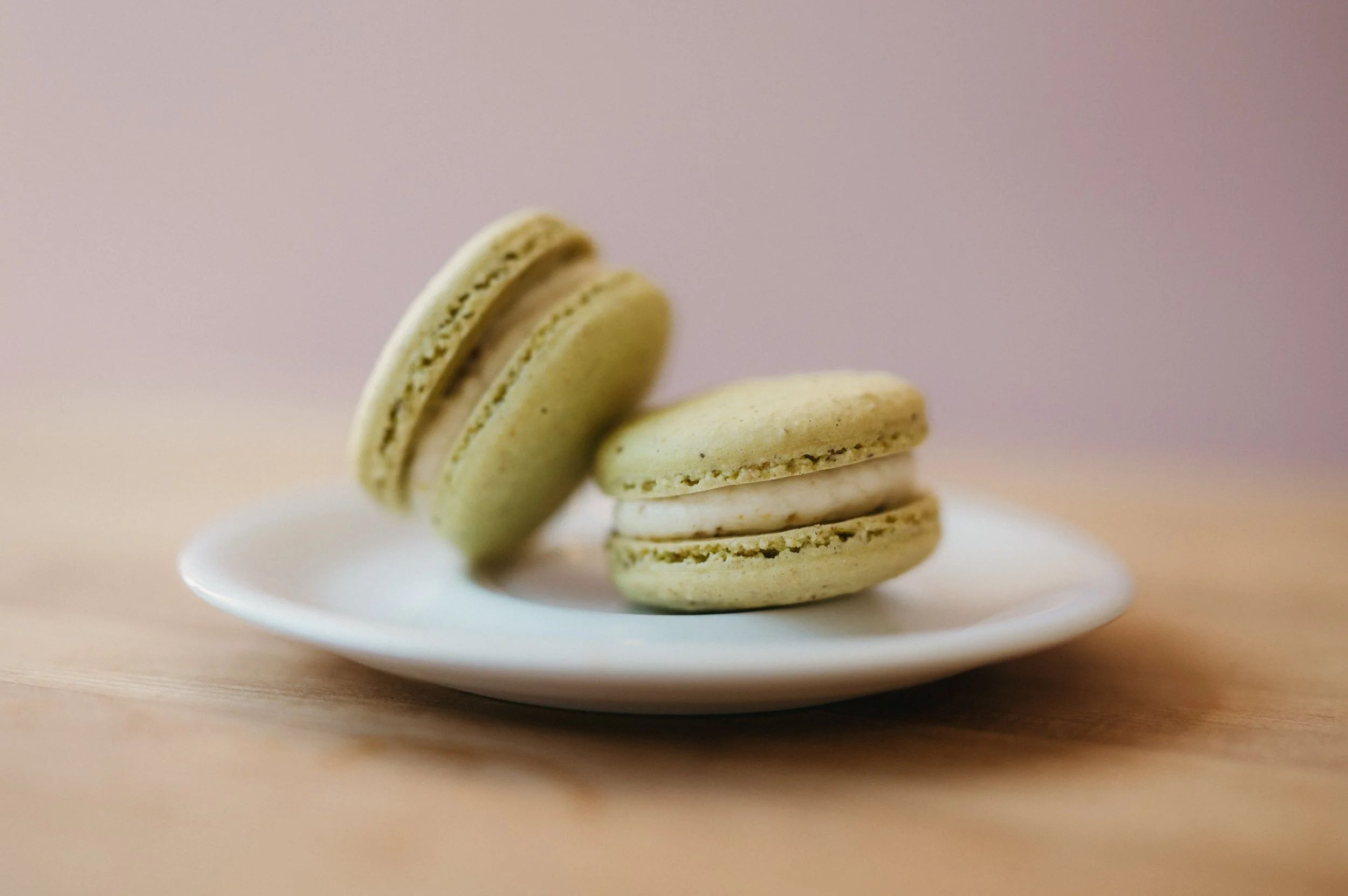 Four macarons, two green, one white, and one beige, stacked on a white plate with a beige background.