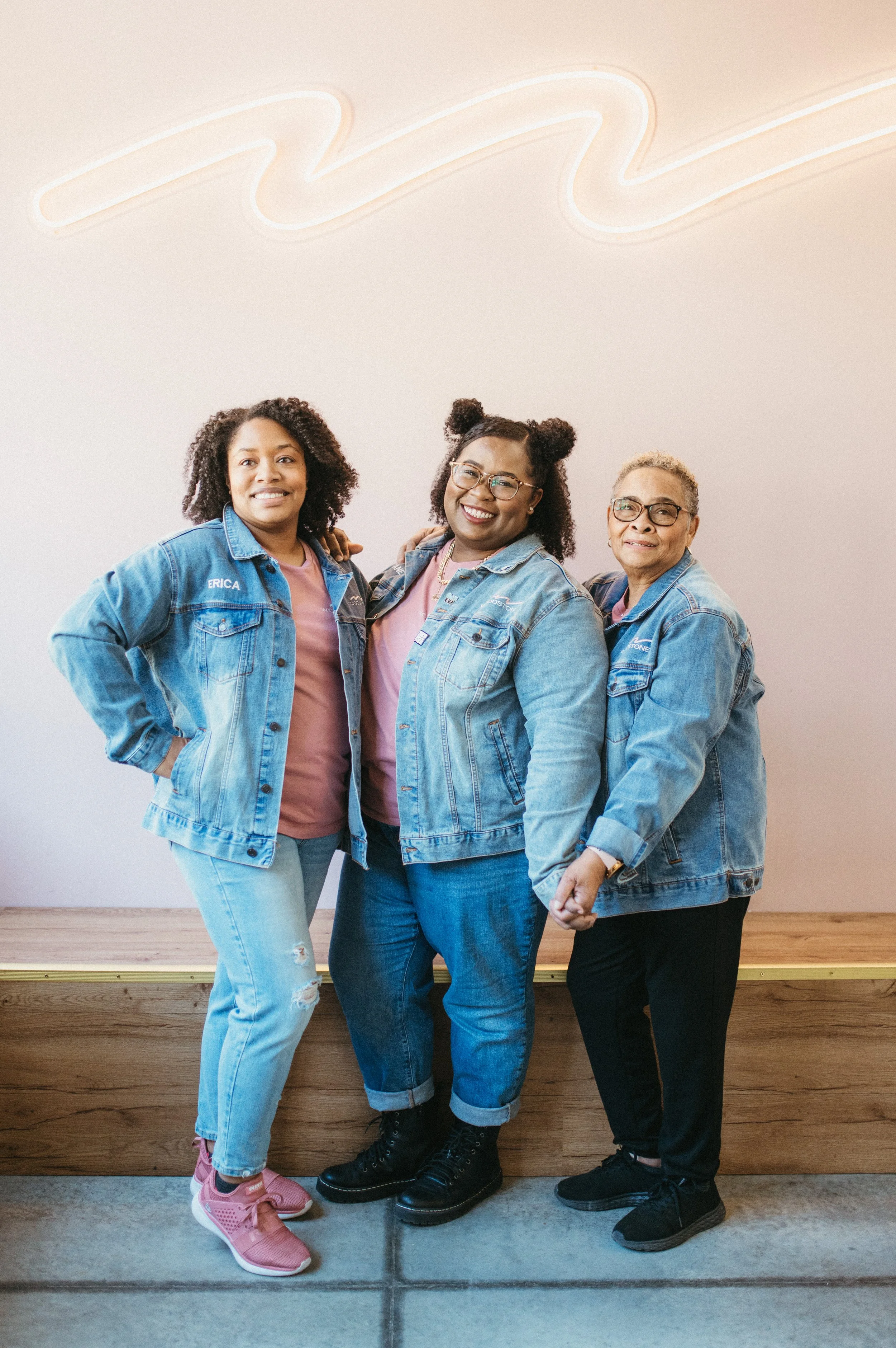 Three women standing together, smiling, wearing denim jackets, and standing in front of a white wall with a neon sign.