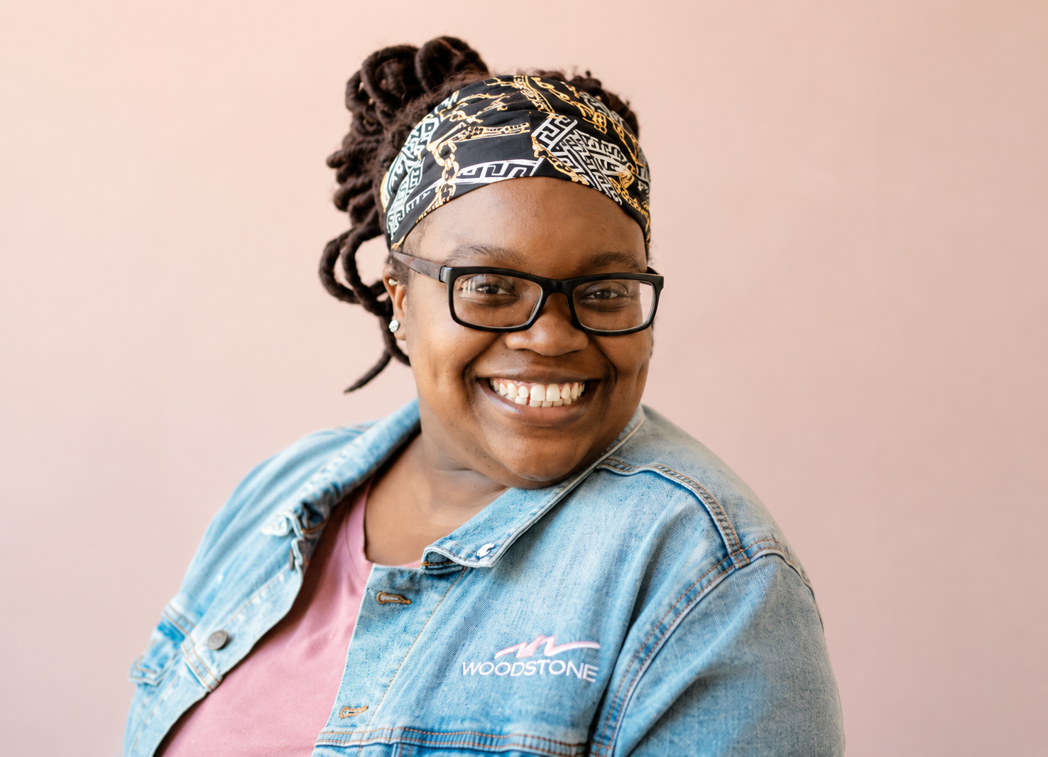 A smiling woman with glasses, wearing a colorful headscarf and denim jacket with the 'Woodstone' logo, poses in front of a light pink background.