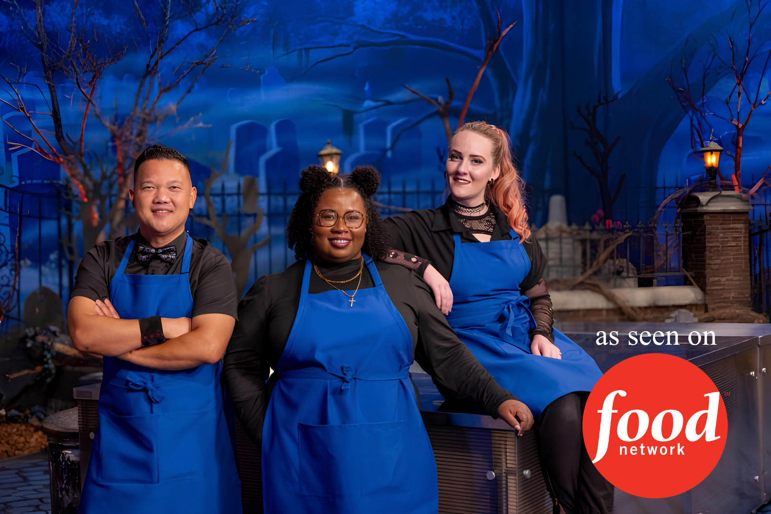 Three people stand in front of a Halloween-themed backdrop wearing blue aprons. The man on the left has short dark hair, the woman in the middle has curly hair, glasses, and is wearing a cross necklace, and the woman on the right has long, light-colored hair. The background features spooky trees, a fence, and a lit lantern. The image has a "Food Network" logo with the text "as seen on" in the lower right corner.