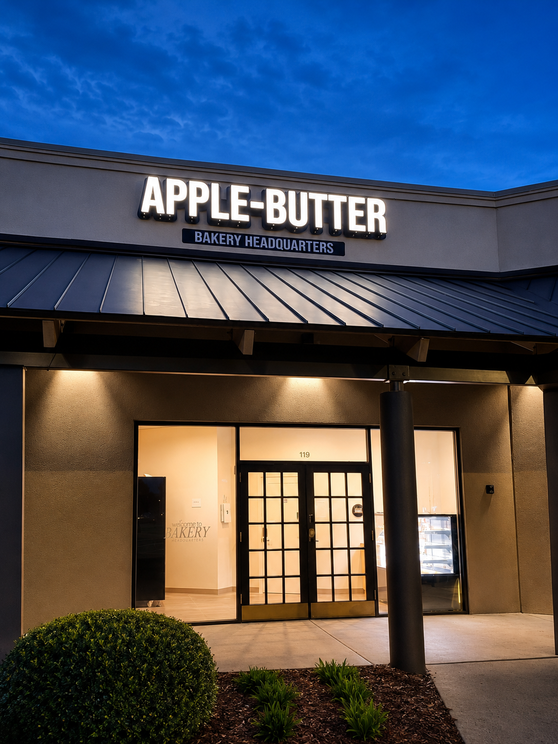 Exterior view of Apple-Butter Bakery Headquarters at dusk with a lit sign, glass doors, and a small garden in front.