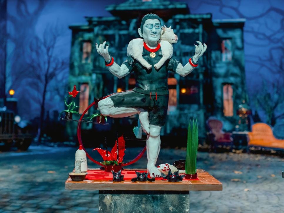 A colorful sculpture of a person with a white dog on their shoulders, standing in a yoga pose with arms raised and eyes closed, in front of a haunted house backdrop.
