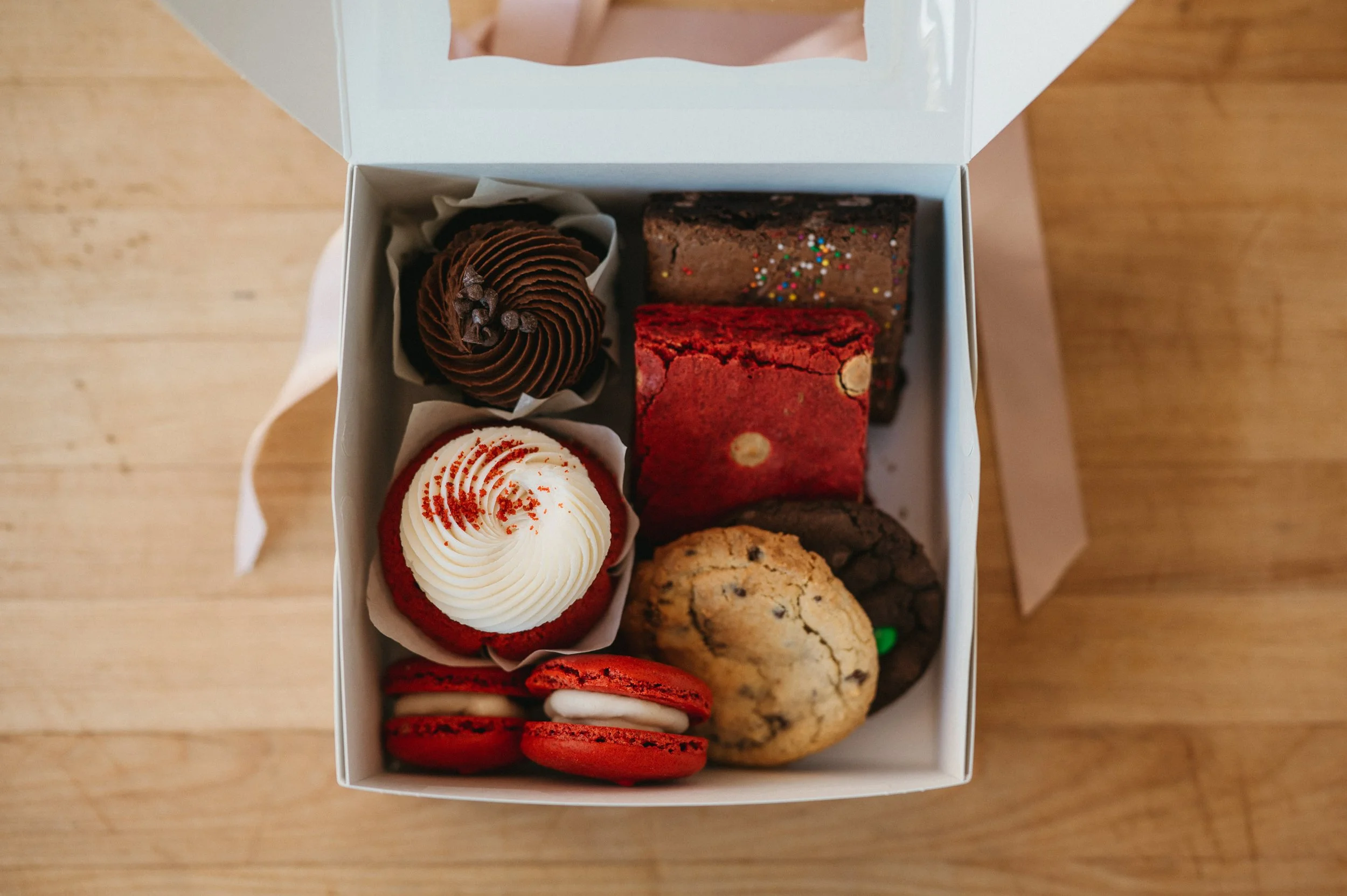 A white box containing assorted baked goods on a wooden surface. The items include decorated cupcakes, brownies with sprinkles, and cookies.