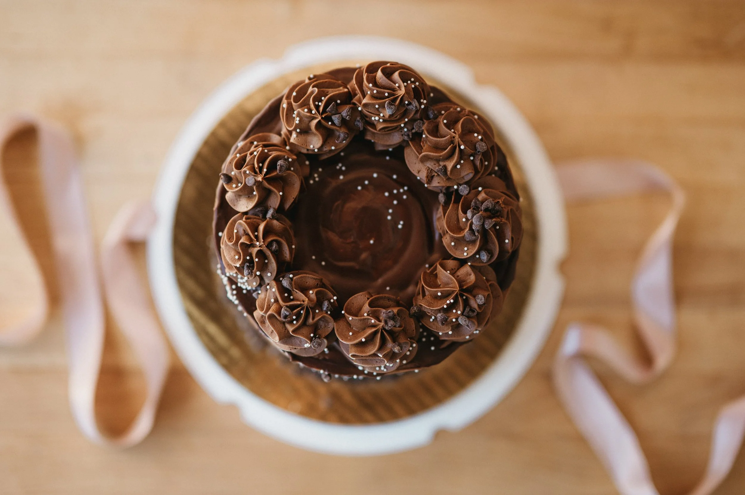 Top view of a chocolate cake decorated with swirls of chocolate frosting, chocolate chips, and white sprinkles, placed in a white paper cup on a wooden surface.