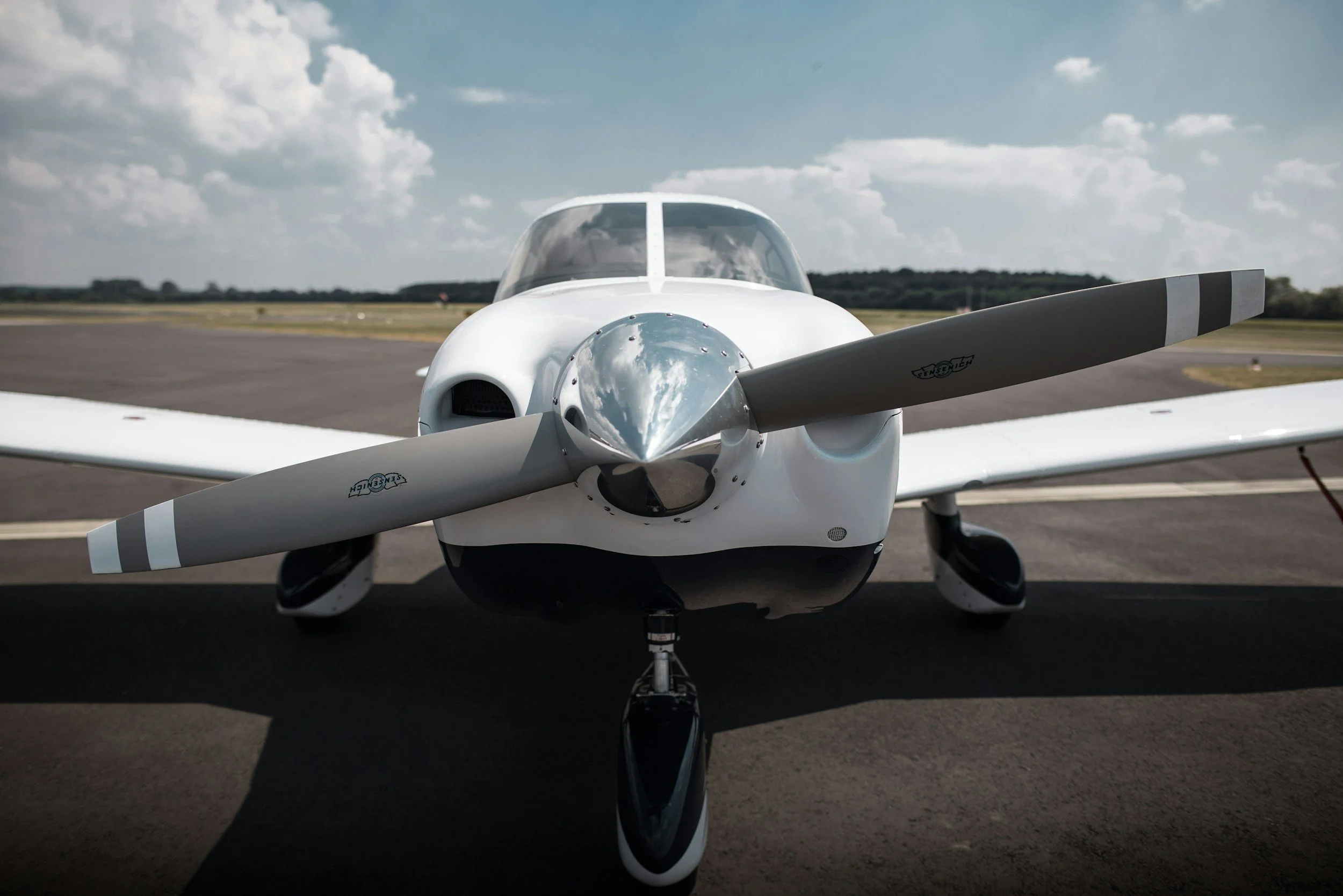 Front view of a small, white, futuristic-looking airplane with a glass cockpit and a propeller on a runway under a partly cloudy sky.