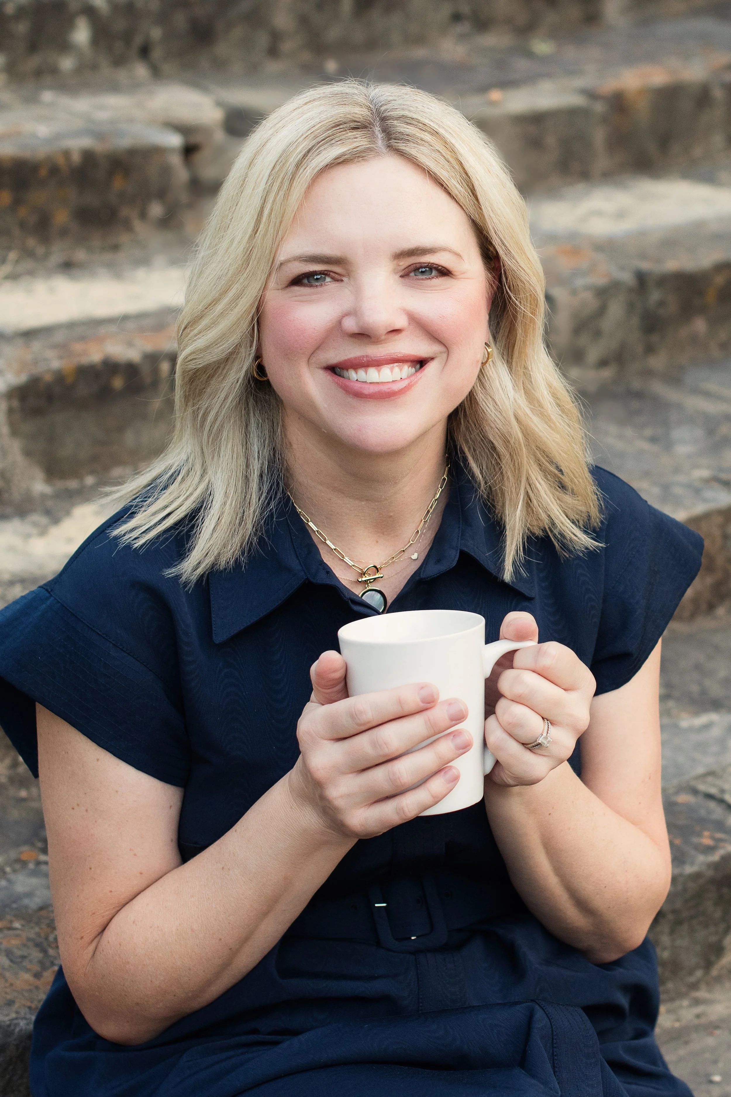 A woman with blonde hair smiling and holding a white mug outdoors with rocks in the background.