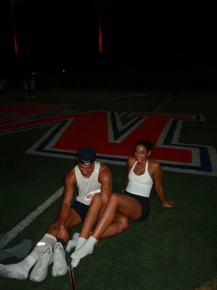 Two athletes, a man and a woman, sit on a sports field at night, smiling and stretching. They are wearing athletic clothing and socks. A large letter and logo are painted on the field surface behind them.