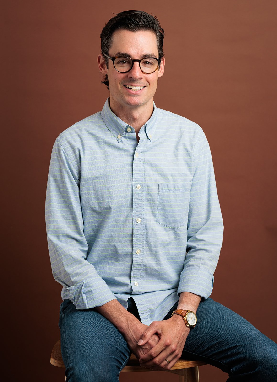 A young man with dark hair wearing glasses and a light blue, striped button-up shirt. He is sitting on a wooden stool against a brown background, smiling with his hands clasped on his lap.