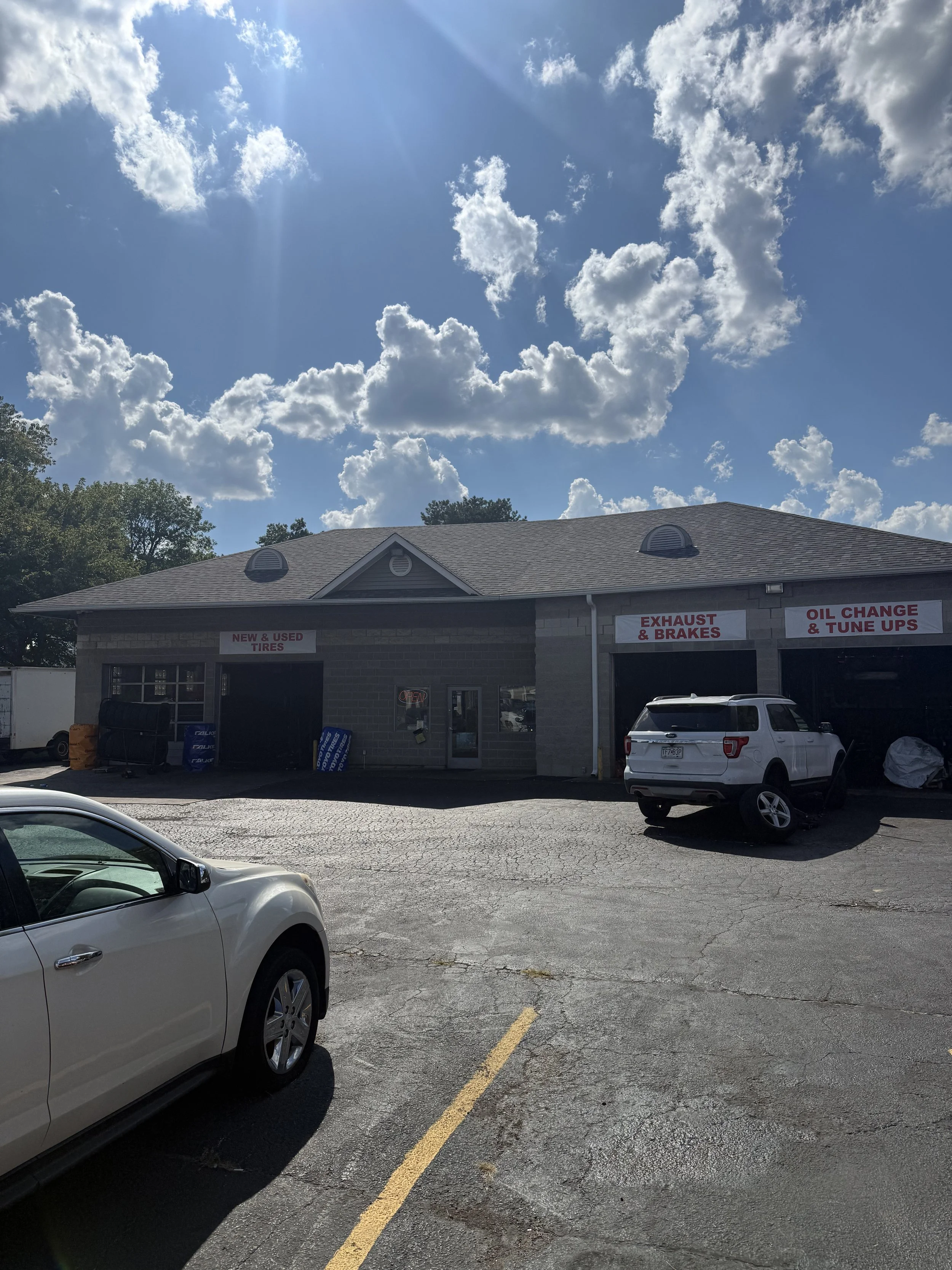 A view of a commercial garage building with signs advertising tire services, exhaust and brakes, oil changes, and tune-ups. The parking lot has two cars visible, and the sky above is bright with scattered clouds.