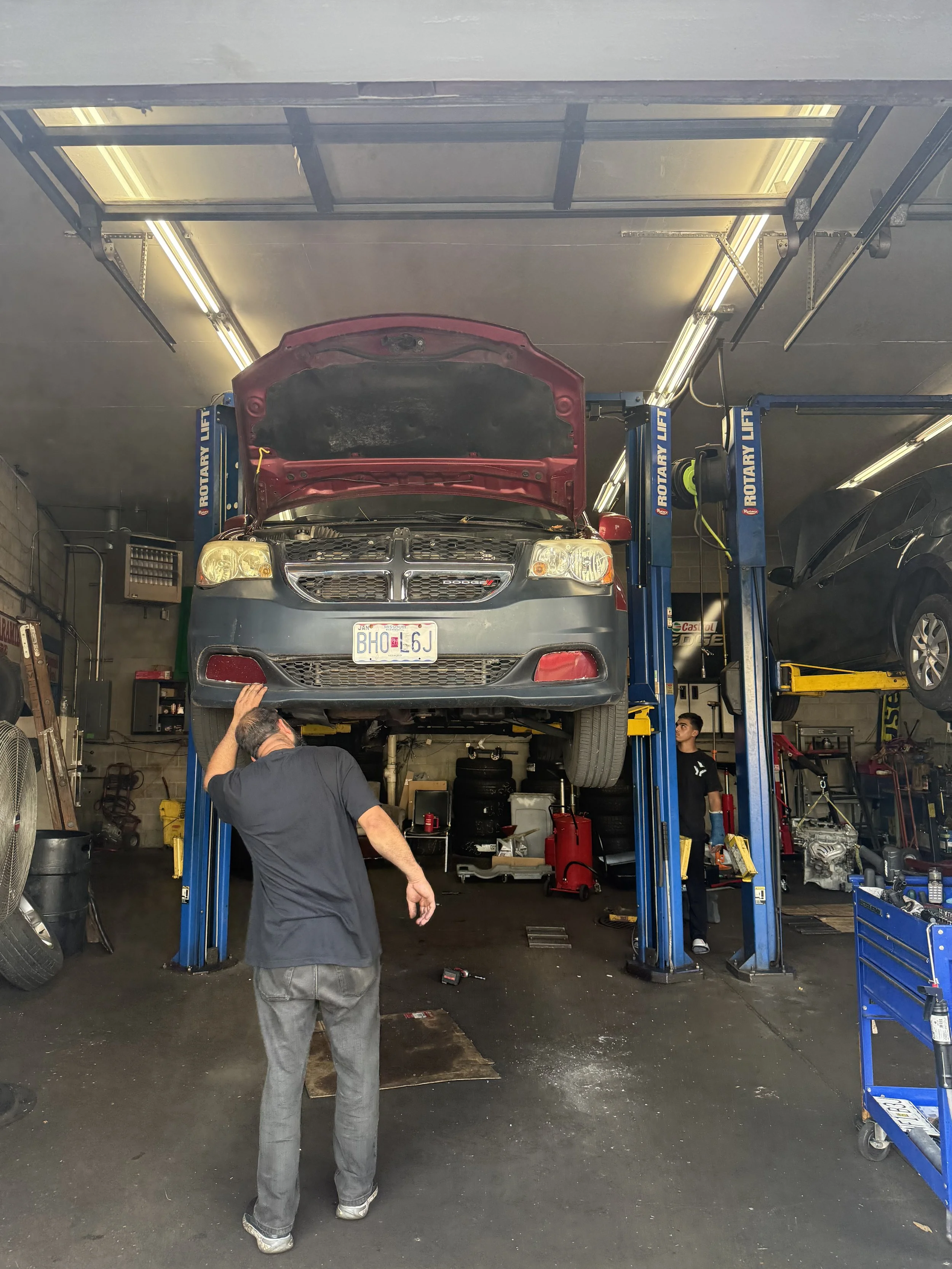 An auto repair shop with a red car on a blue lift. Two mechanics are working; one is inspecting beneath the car, the other stands nearby. The shop has tools and equipment, and fluorescent lights on the ceiling.