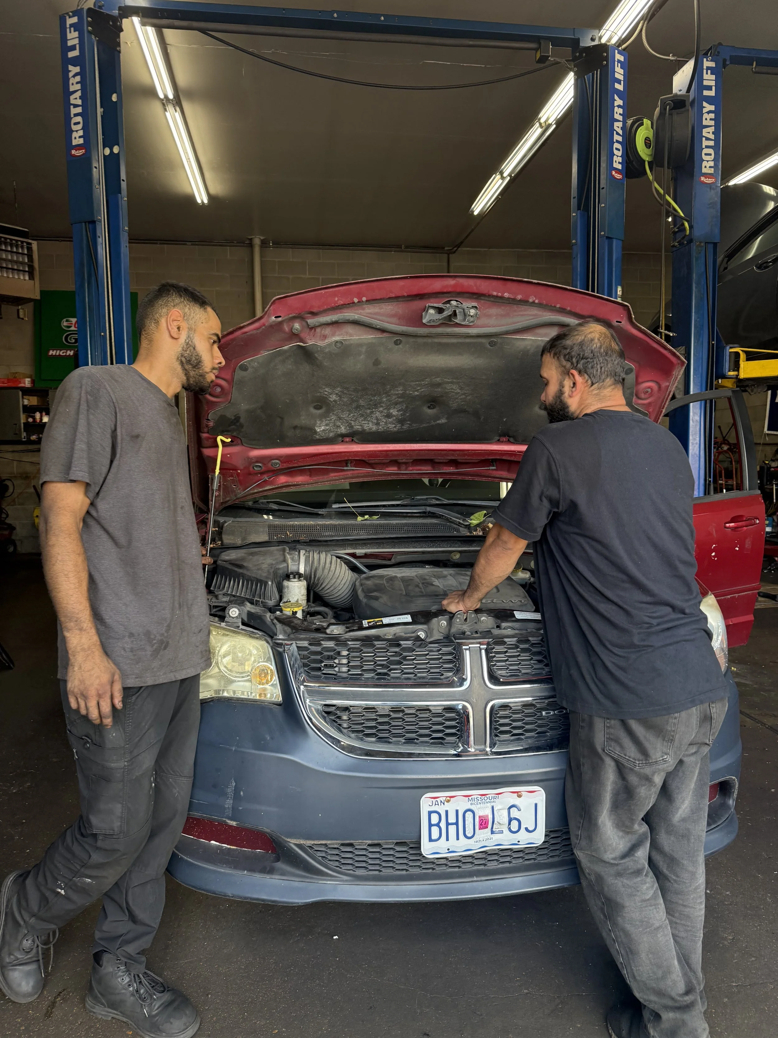 Two men working on or inspecting a car engine in a garage, with the car's hood open.