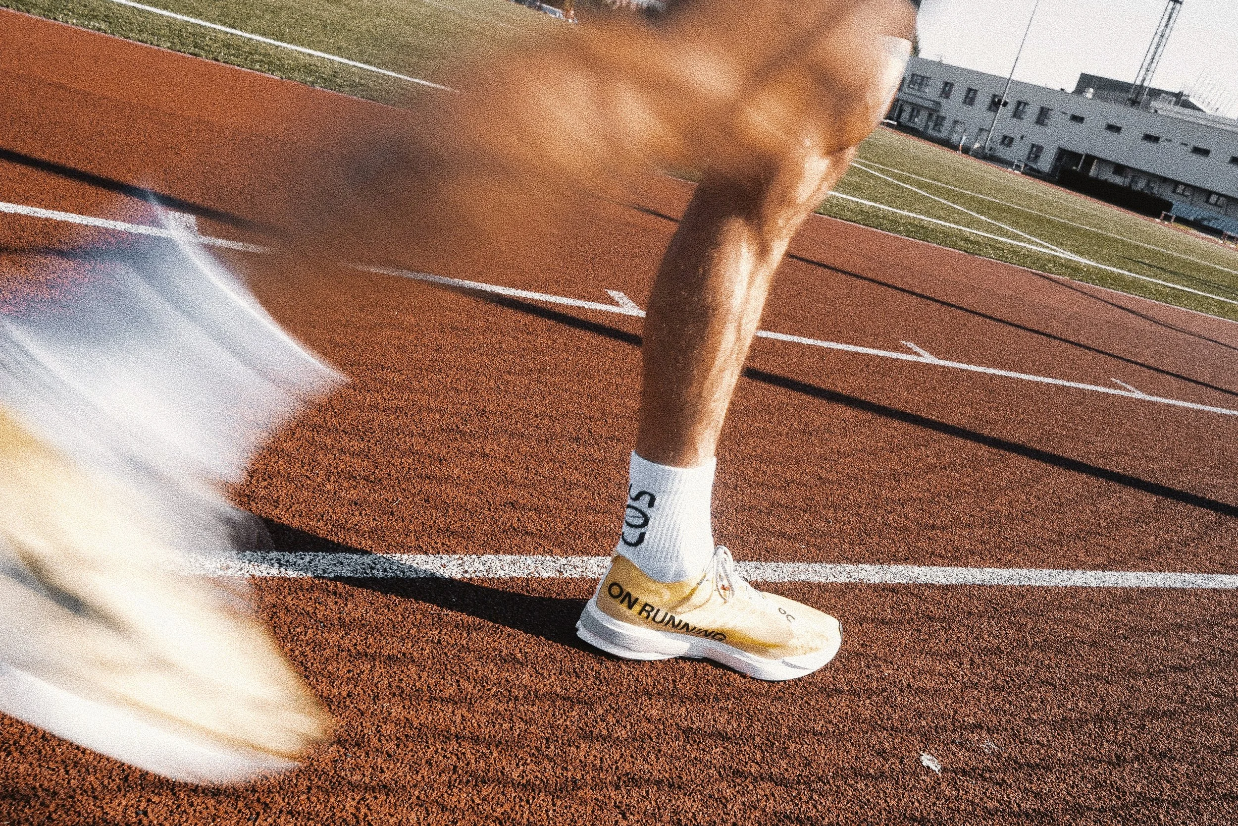 A close-up of a runner's legs on a red running track. The runner is wearing white running shoes with yellow accents and white socks that have text on them. The background shows the running lanes and some buildings in the distance.