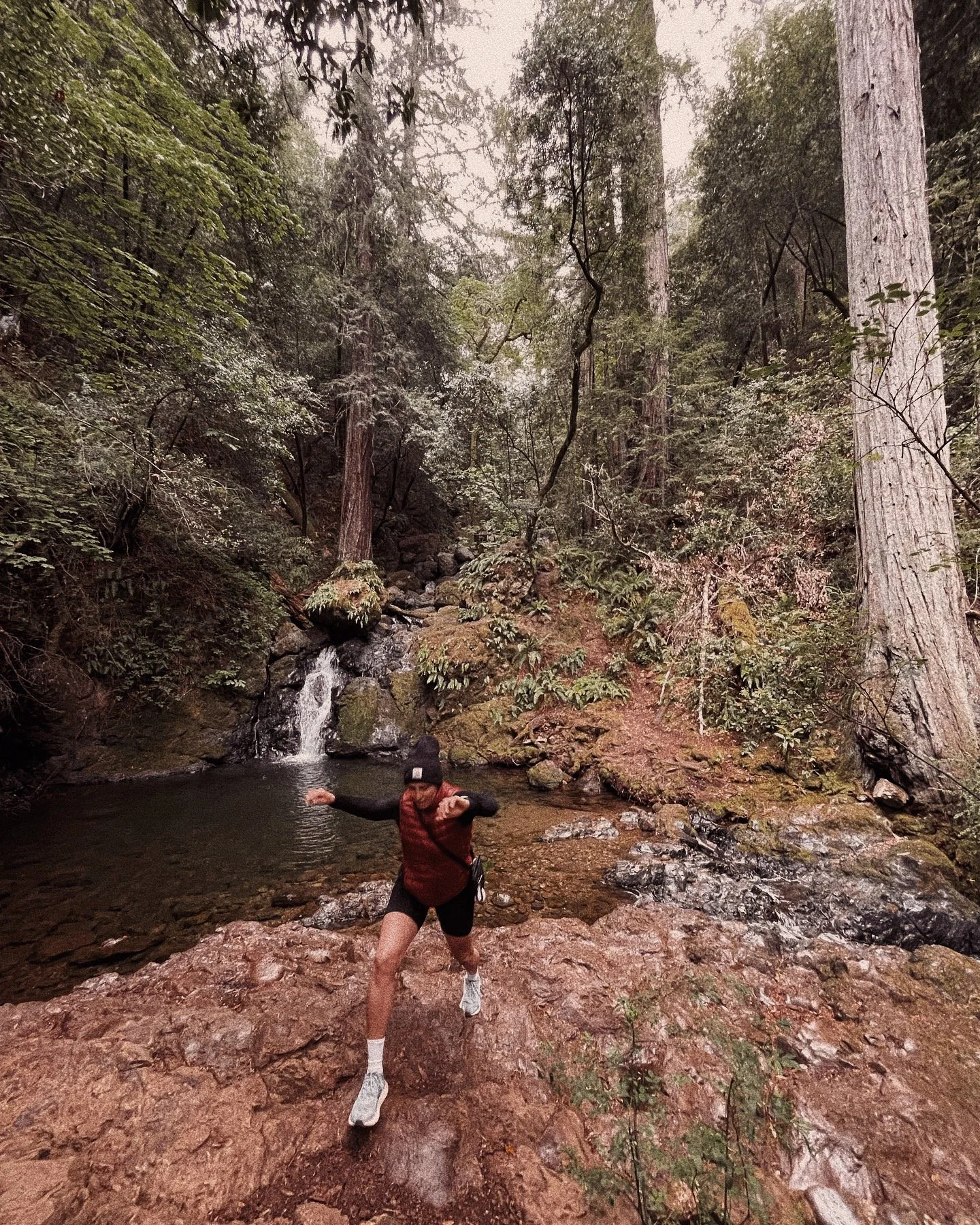 A person standing in a shallow stream or river in a forest with tall trees and a small waterfall overhead, wearing a red vest, black shorts, a black cap, and white shoes, splashing water.