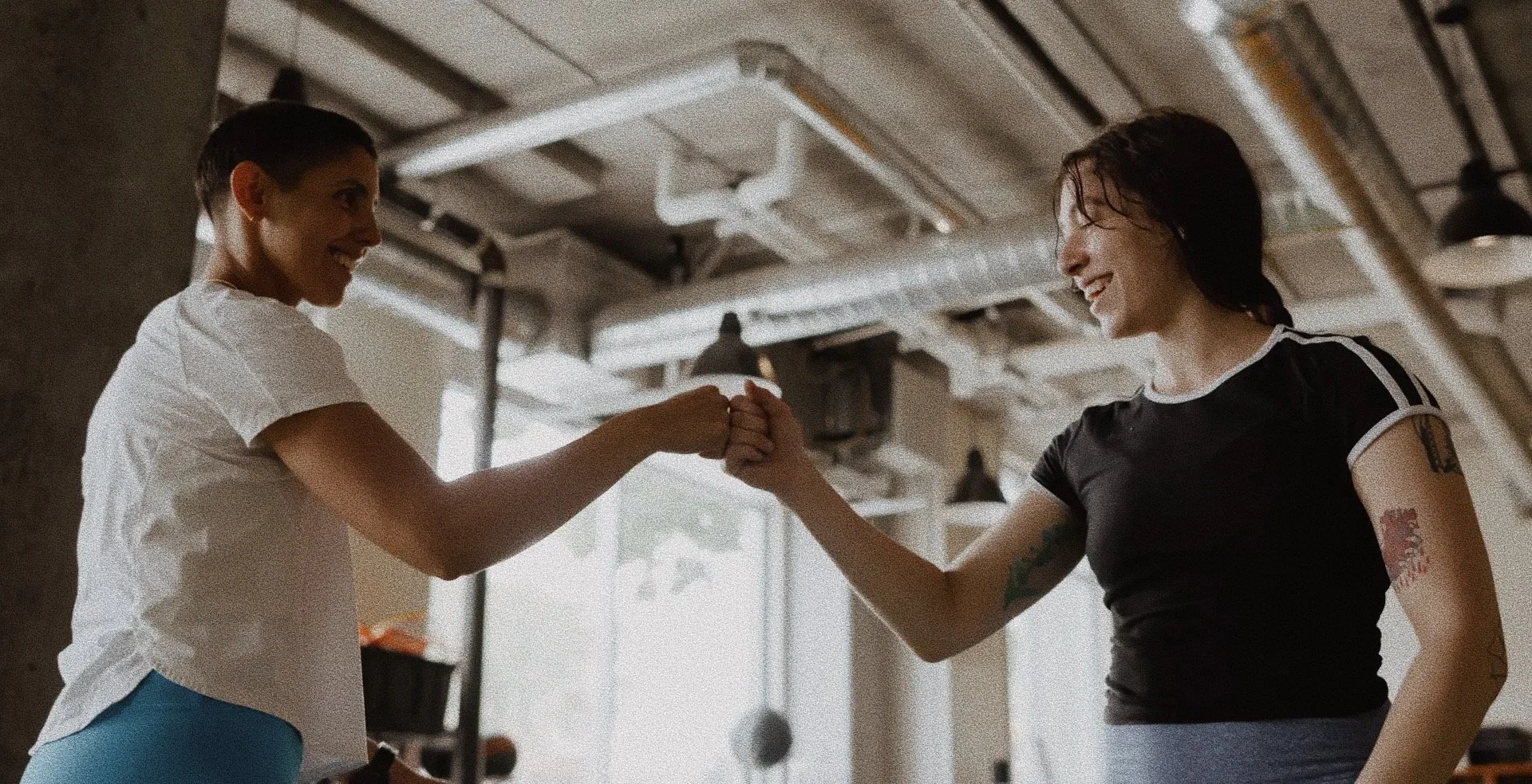 Two women smiling and fist bumping inside a gym with a ceiling fan and industrial lighting.