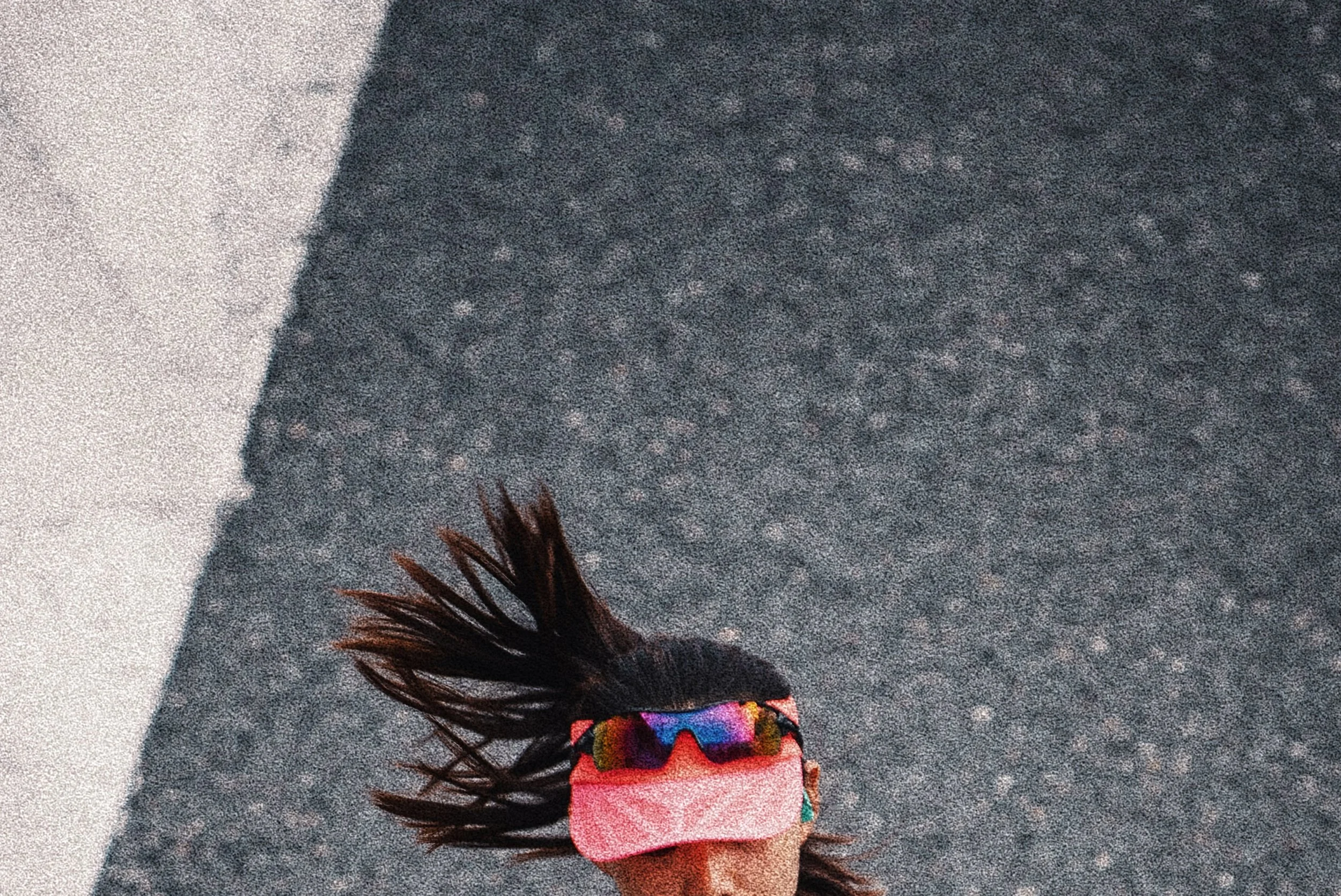 A person standing on a gray carpeted floor with long dark hair, wearing rainbow-colored glasses, a multicolored shirt, and a pink wristband.
