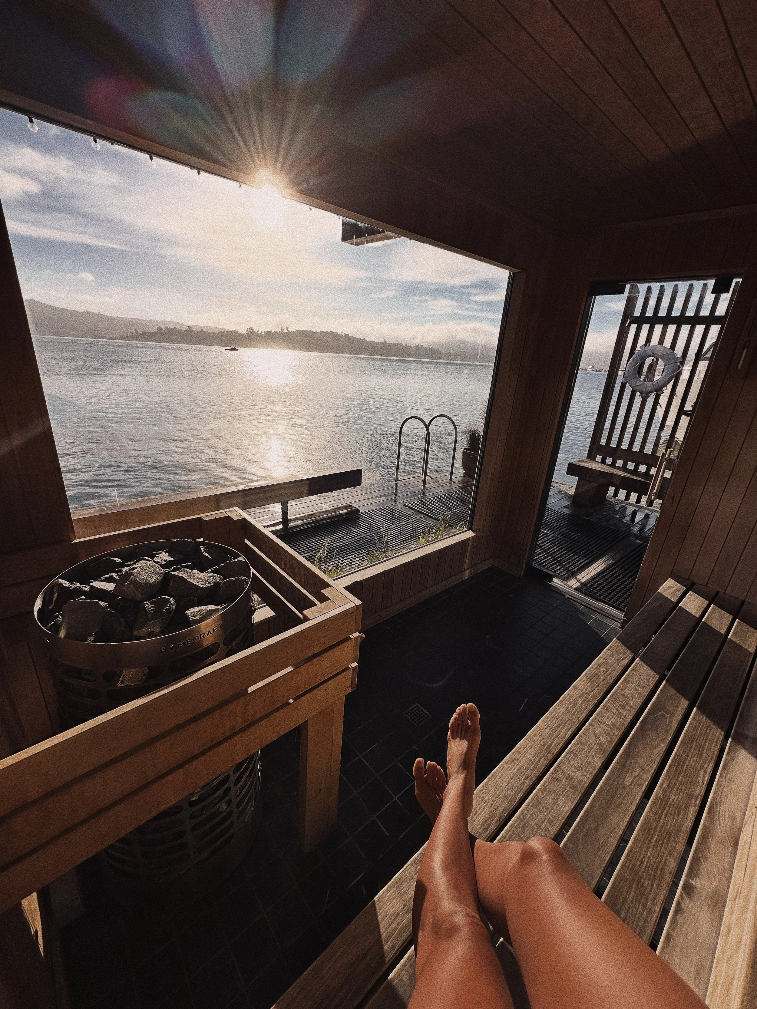 View from inside a sauna or steam room, showing a person's legs and feet resting on wooden benches, with a bucket of stones and a sauna heater visible, overlooking a body of water and distant hills under a bright sky.