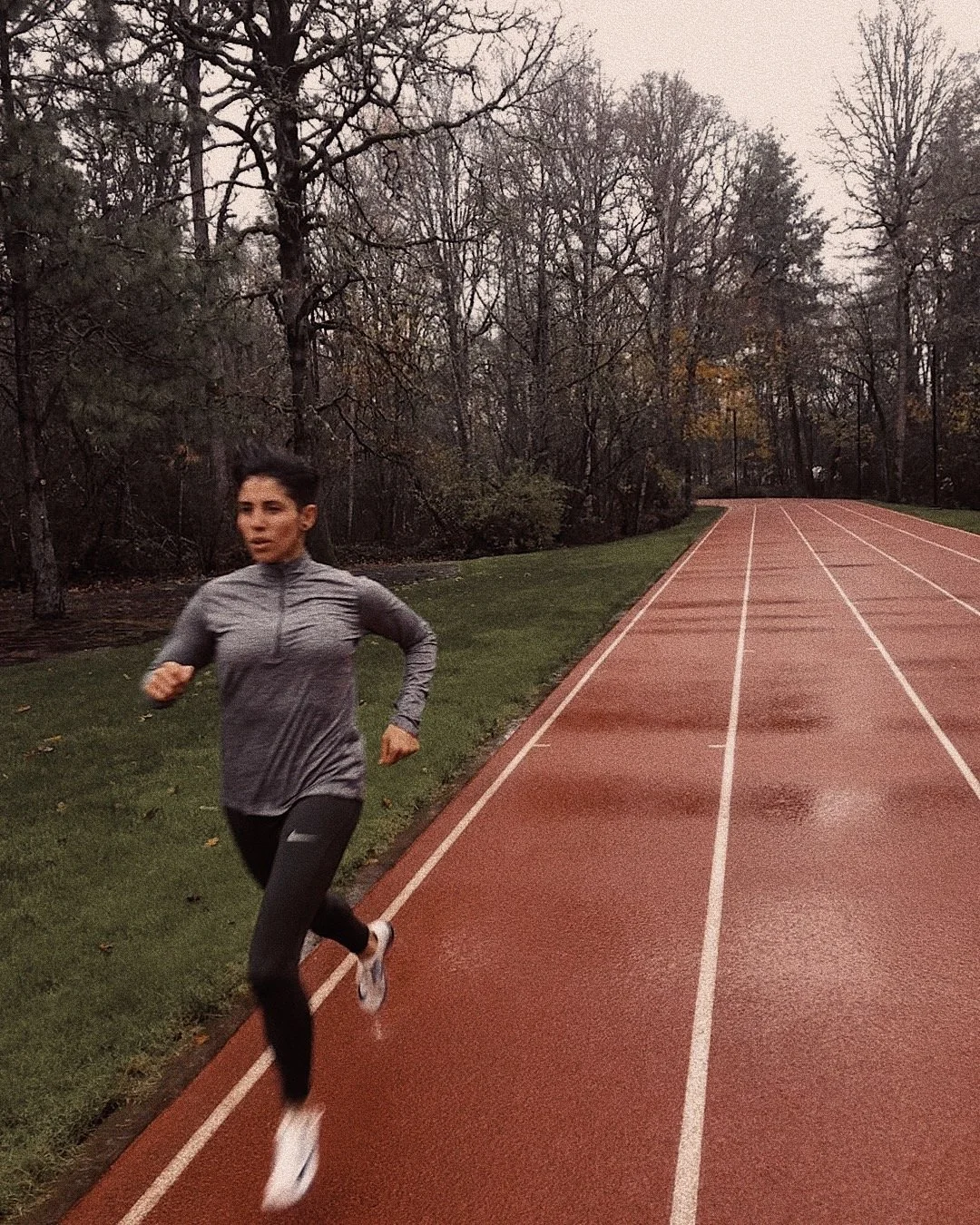 A woman running on an outdoor track surrounded by trees during a cloudy day.