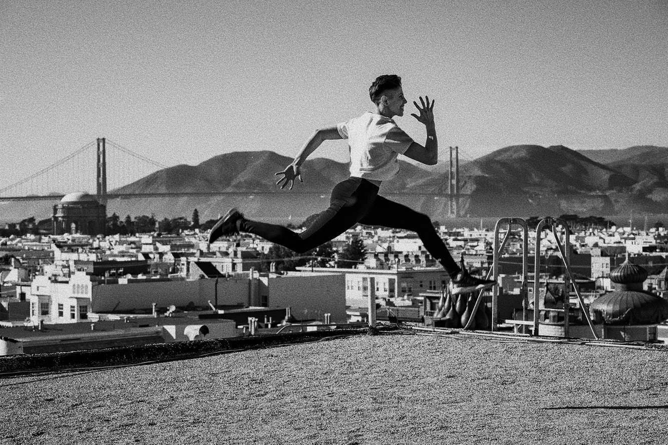 A person is leaping across a rooftop in a cityscape with hills and bridges in the background. The scene is in black and white.