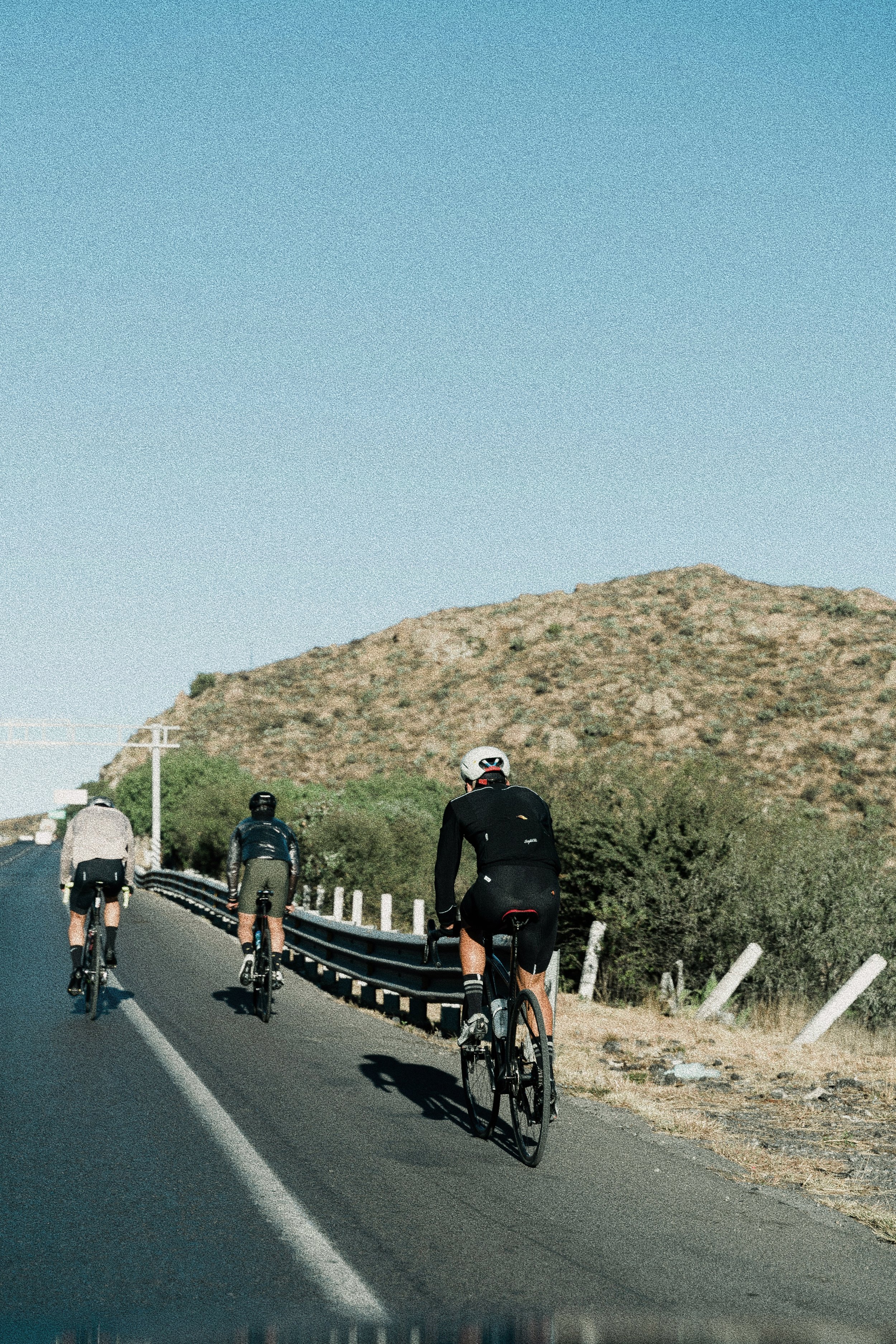 A group of five cyclists riding on a winding road through a hilly landscape with dry, sparse vegetation and a clear blue sky.