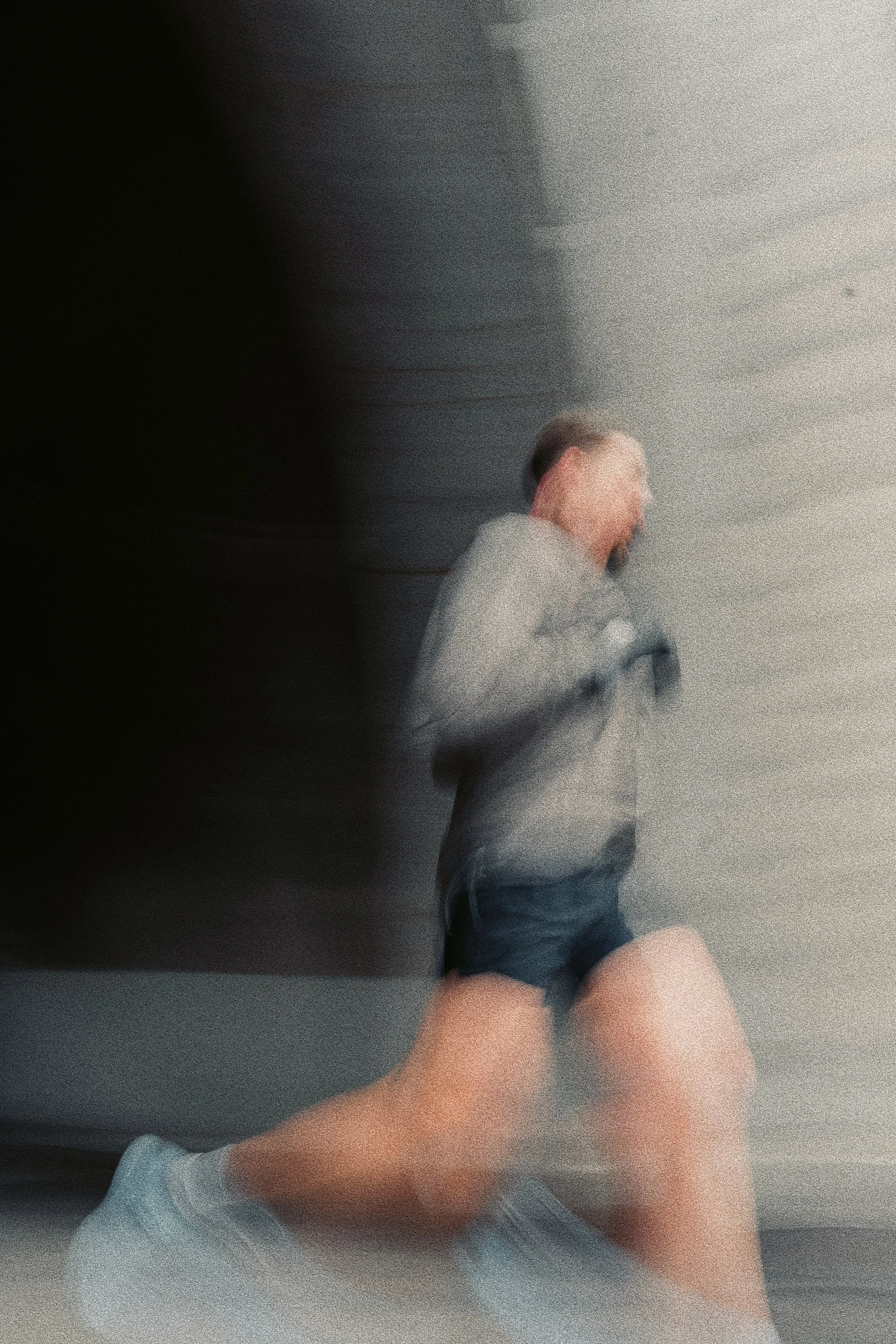 Blurred photo of a person with short hair, wearing a gray shirt and dark shorts, standing in front of a wall.