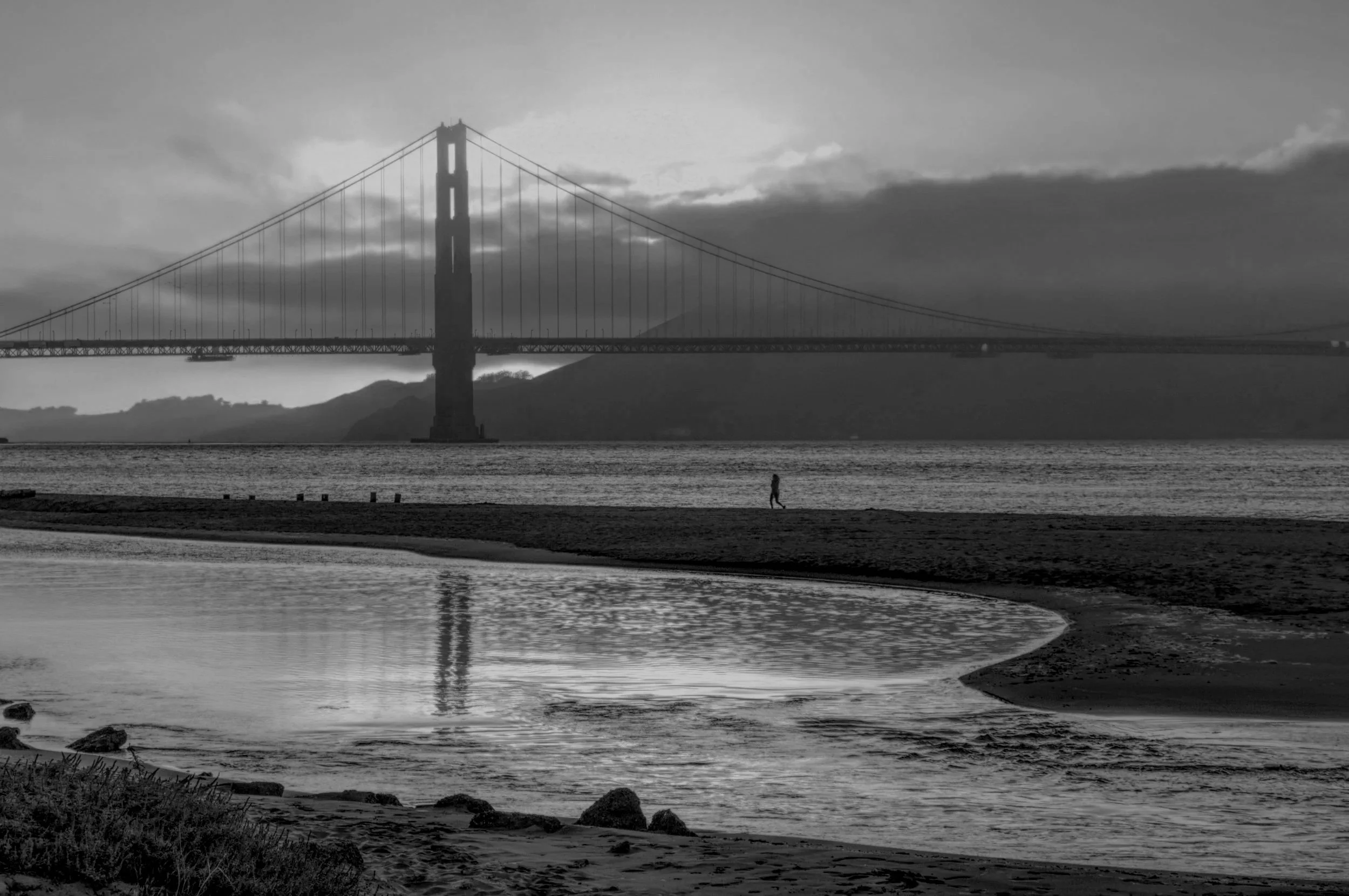 Black and white photo of the Golden Gate Bridge in San Francisco with a person walking along the shore in the foreground, water and hills in the background.