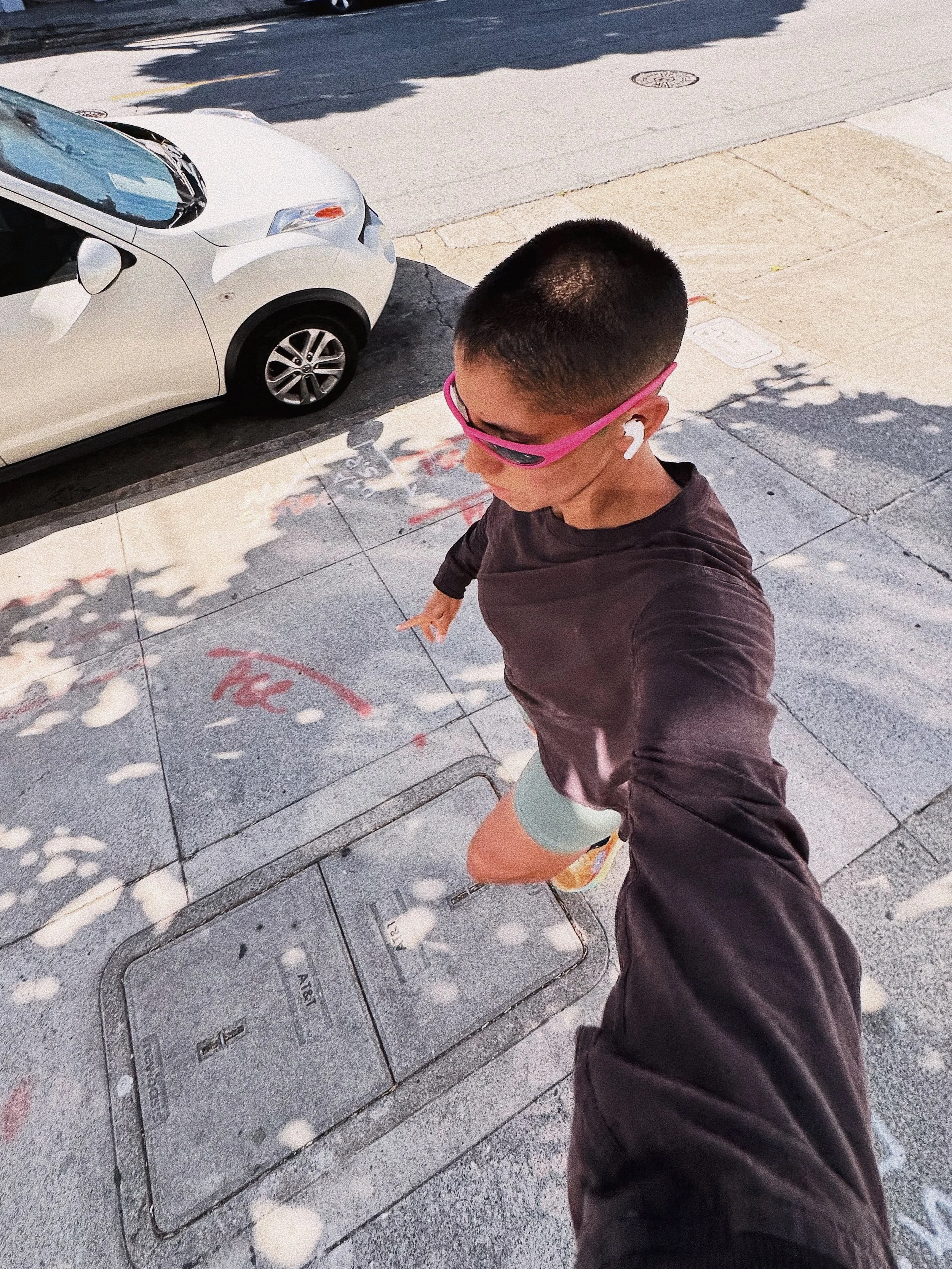A person taking a selfie outdoors, wearing pink sunglasses, outdoor clothing, and earbuds, with a parked white car and sidewalk in the background.