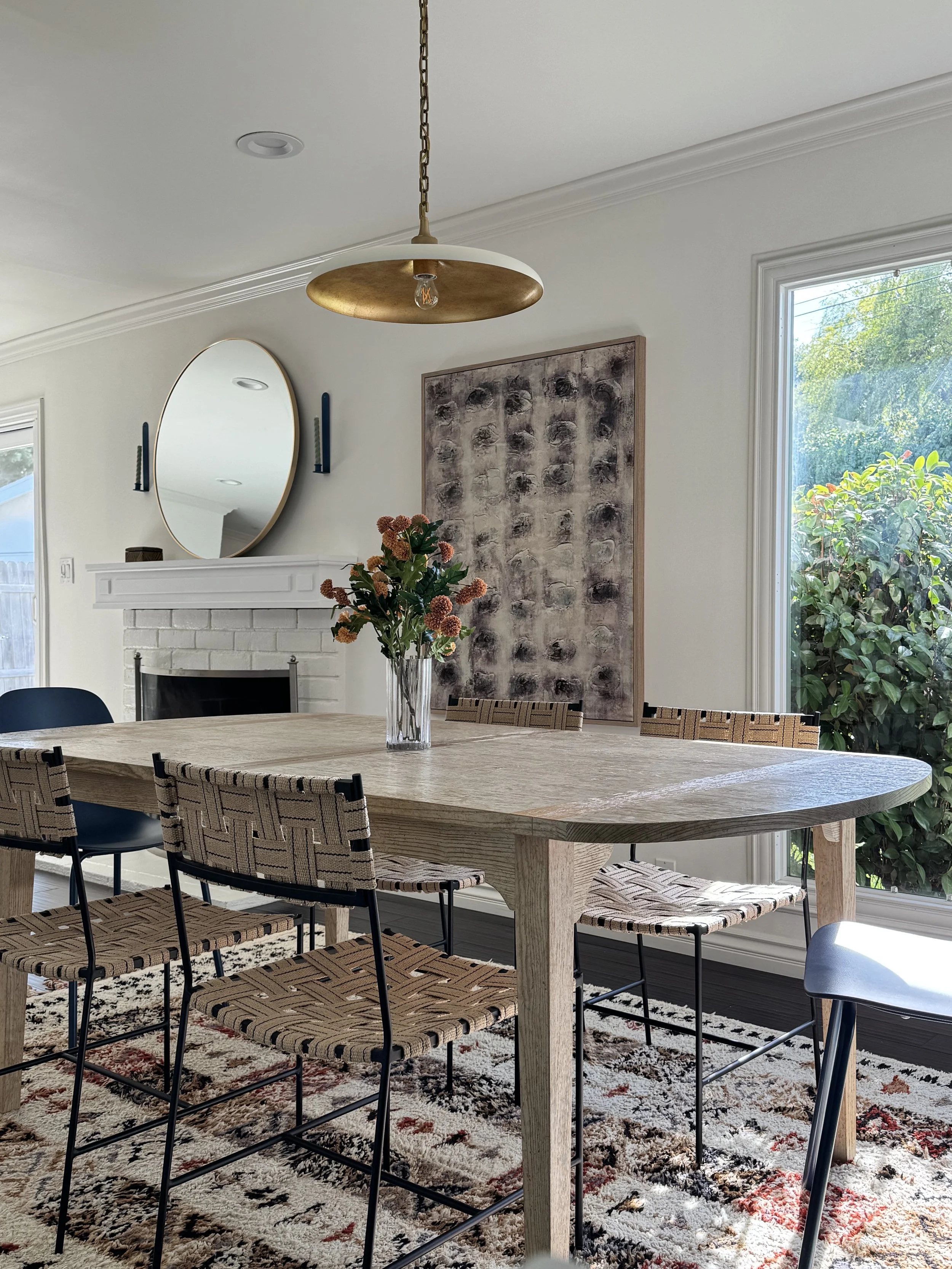 A dining area with a large wooden table, wicker chairs, and a colorful patterned rug. A glass vase with orange flowers is on the table. In the background, there is a white brick fireplace, a round mirror, a large abstract artwork, and windows showing greenery outside. A ceiling light fixture hangs above the table.