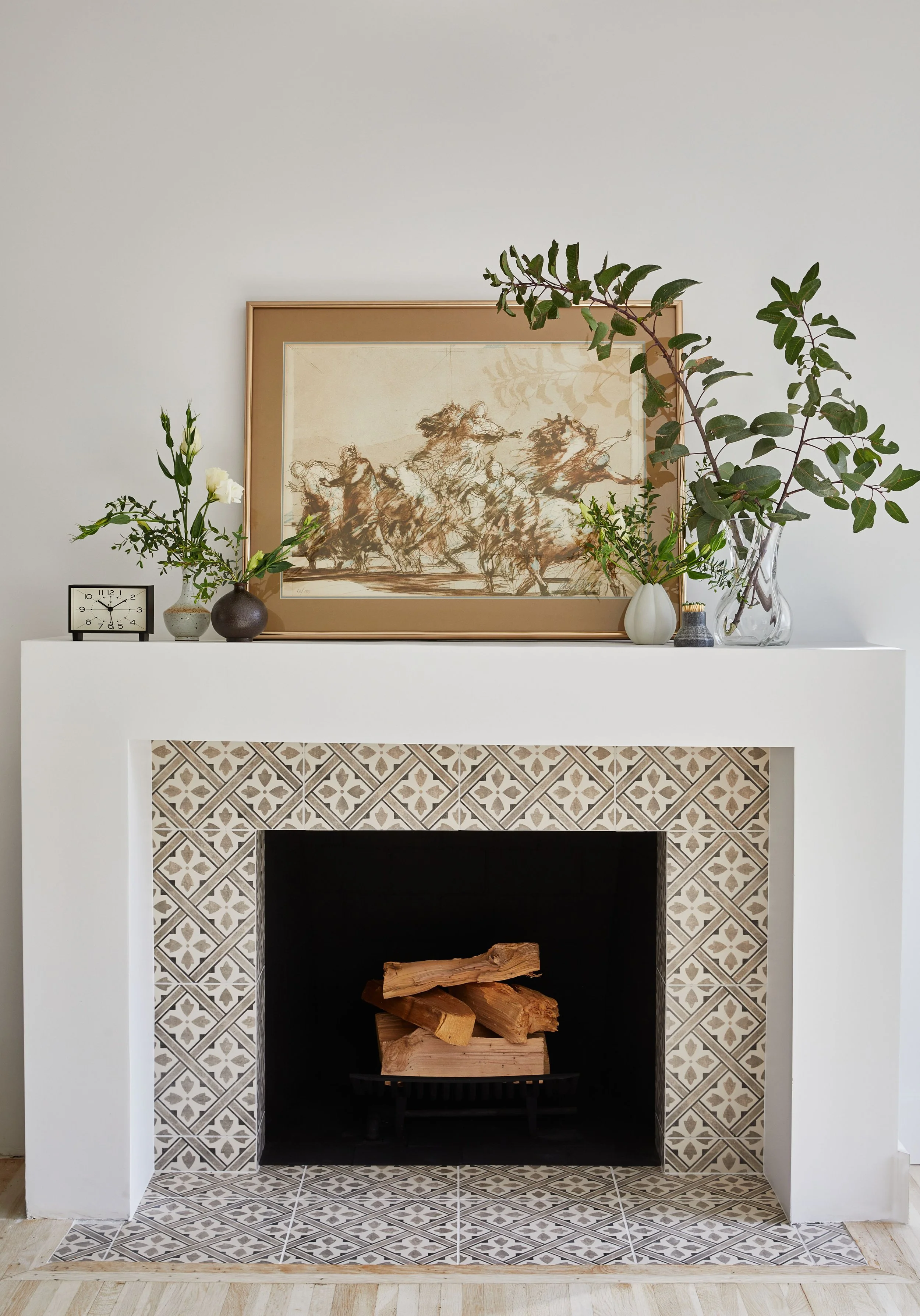 Fireplace with decorative tile surround, stacked firewood inside, and decorative items on mantel including a clock, vases with green foliage, and framed art above.