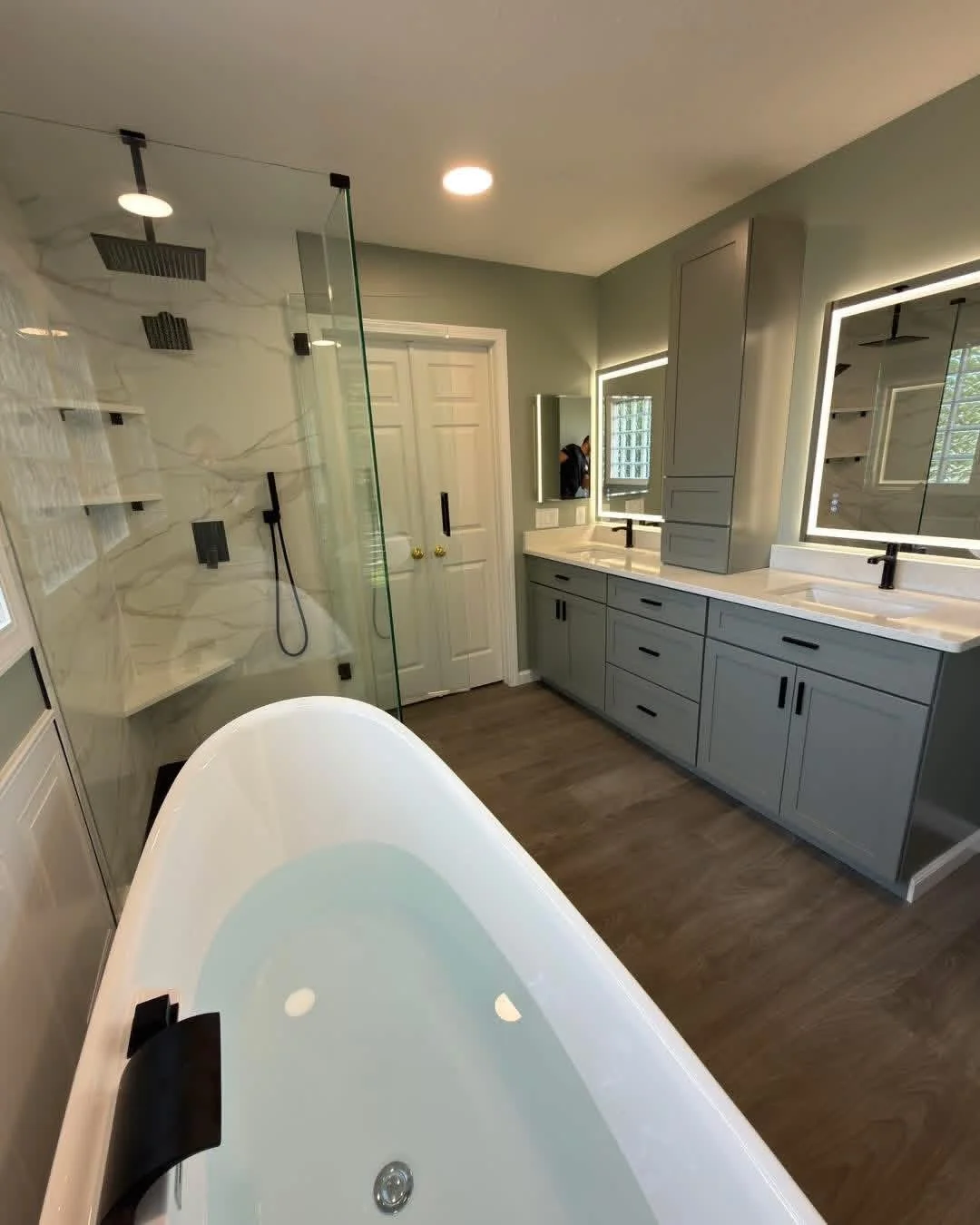 Modern bathroom with bathtub, dual sinks, gray cabinets, large mirrors with LED lights, and a separate glass-enclosed shower with marble walls.
