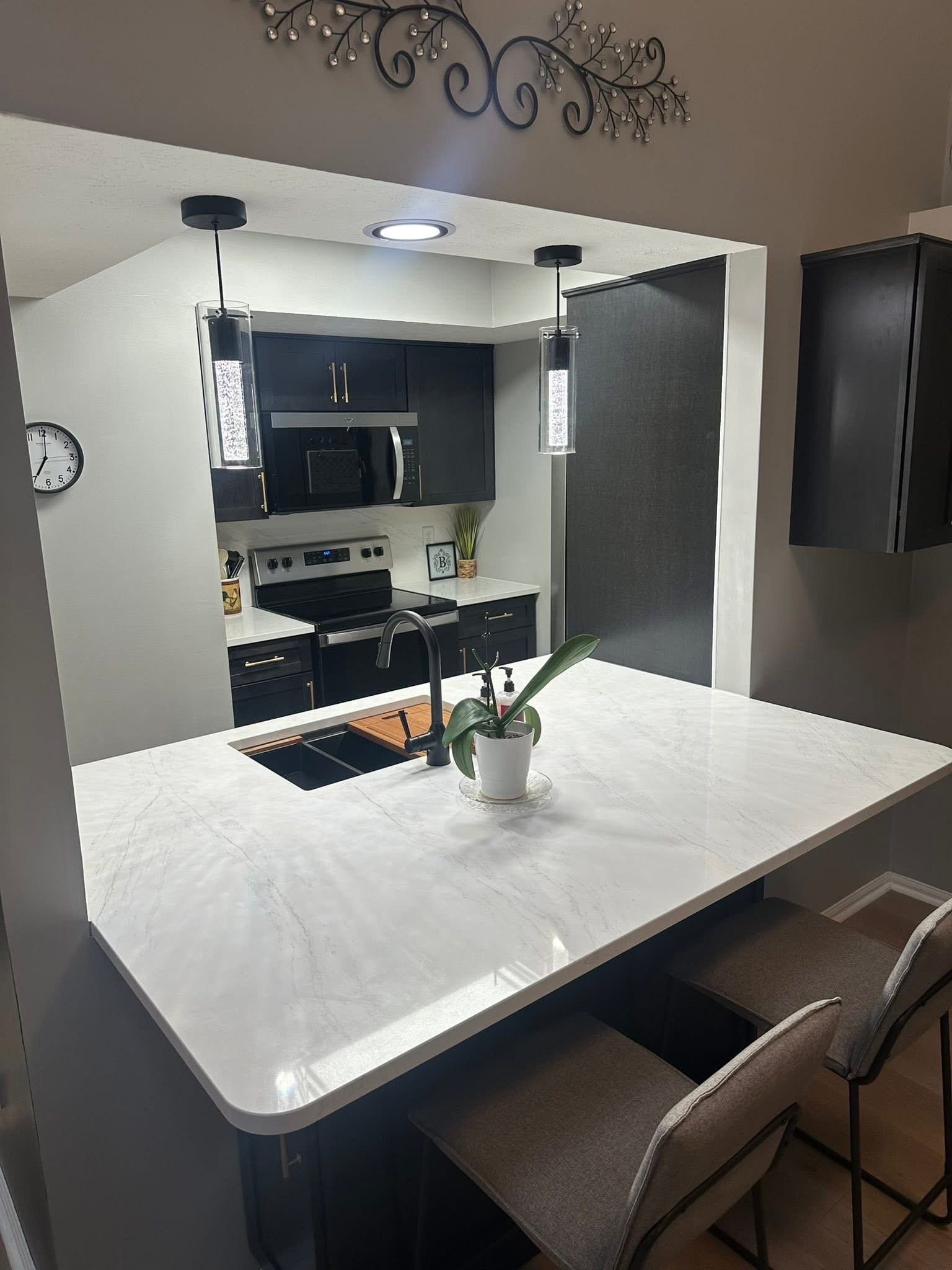 A kitchen with a white marble countertop, dark cabinets, a microwave oven, and a stainless steel stove. There is a plant in a white pot on the counter, a clock on the wall, and two pendant lights hanging from the ceiling.