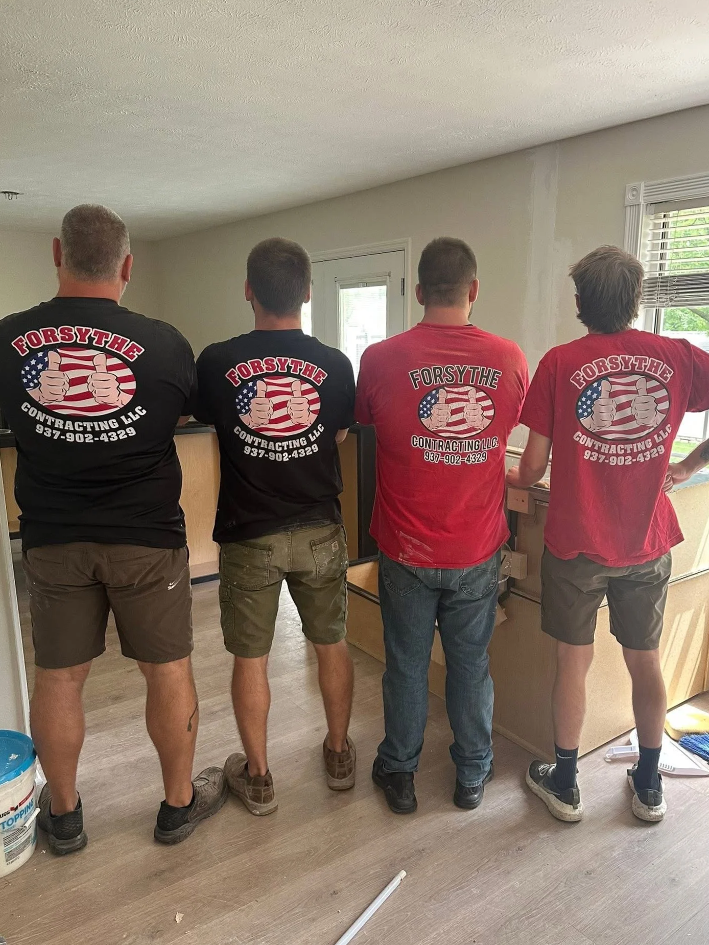 Four men with their backs to the camera, standing indoors, wearing matching t-shirts with 'Forsythe Contracting LLC' logo and phone number, facing a wall with windows.