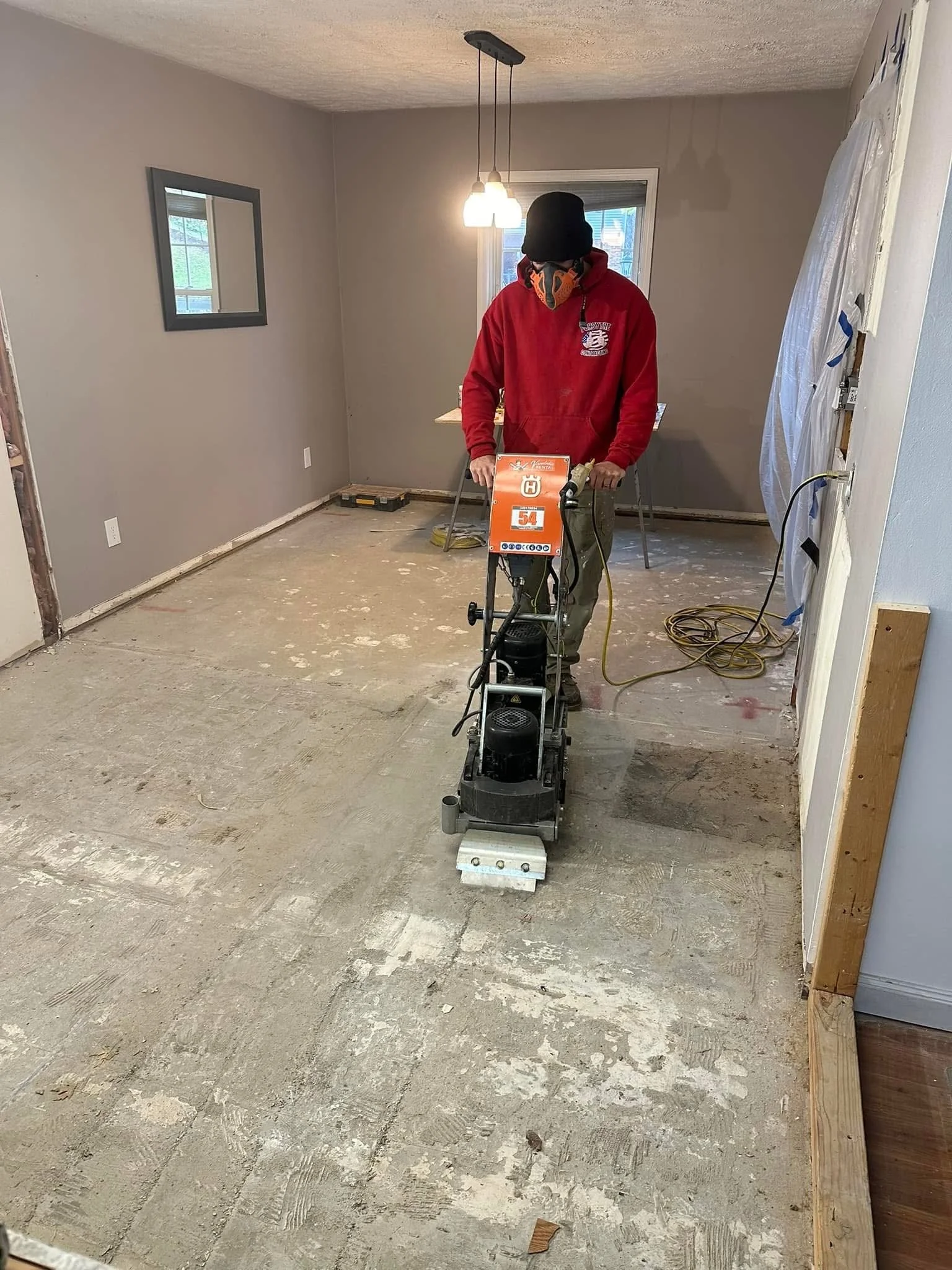 A person wearing a red sweatshirt, black hat, and face mask is operating a floor sander in an unfinished room with gray walls and a concrete floor.