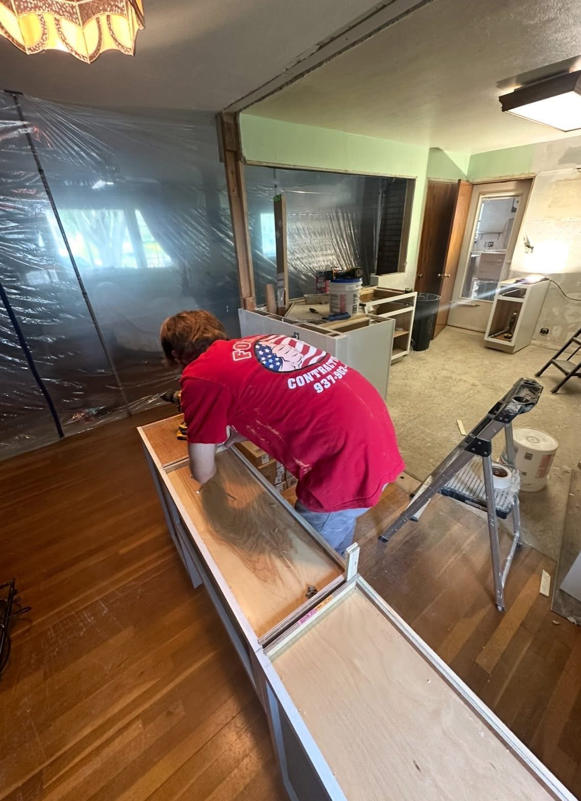 A person installing or repairing a wooden cabinet in a house during renovation, with construction tools, ladder, and plastic sheets covered in the background.