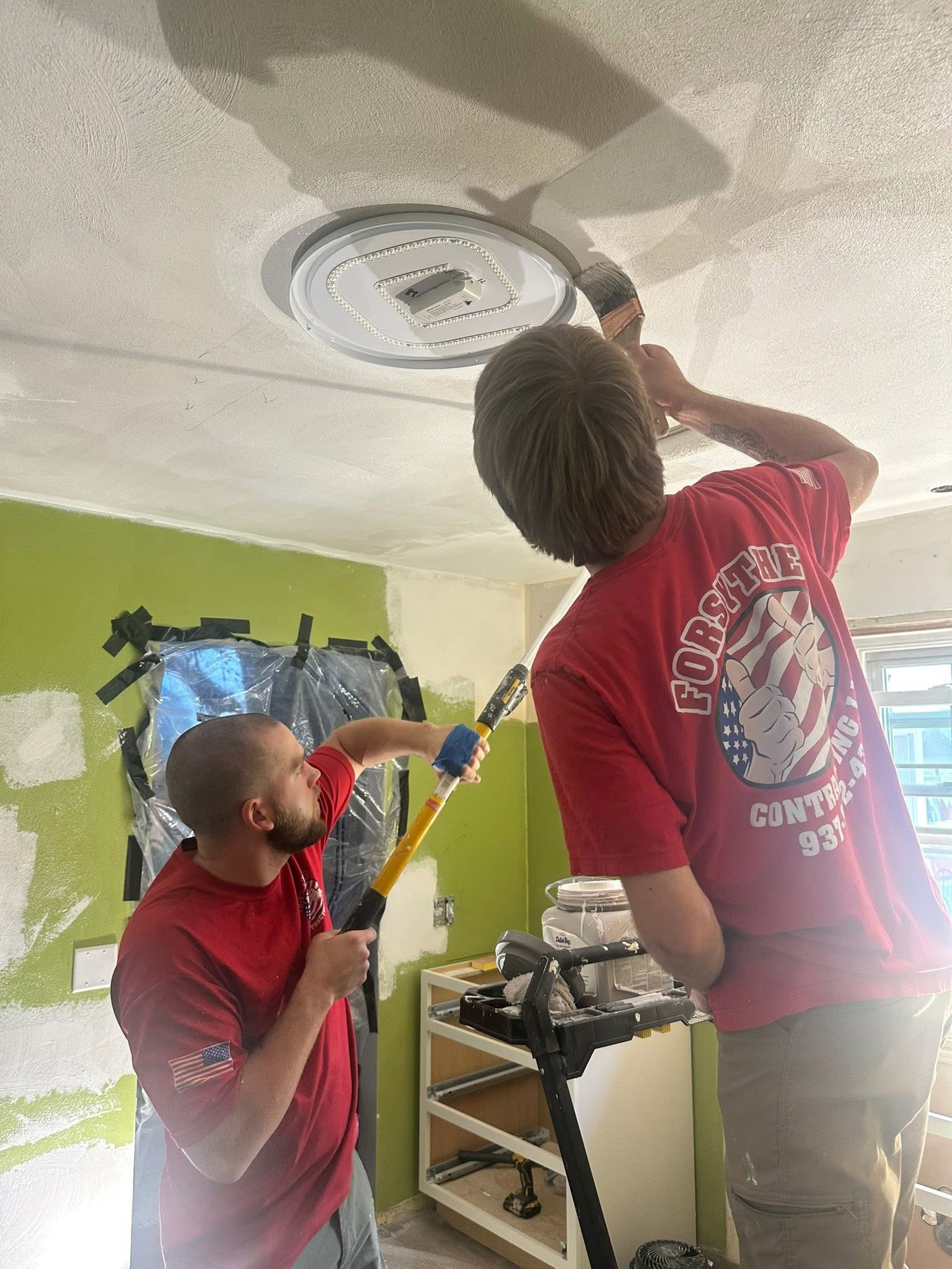Two men painting a ceiling in a room with lime green walls, using paint rollers, while one of them operates an extension pole to reach higher.