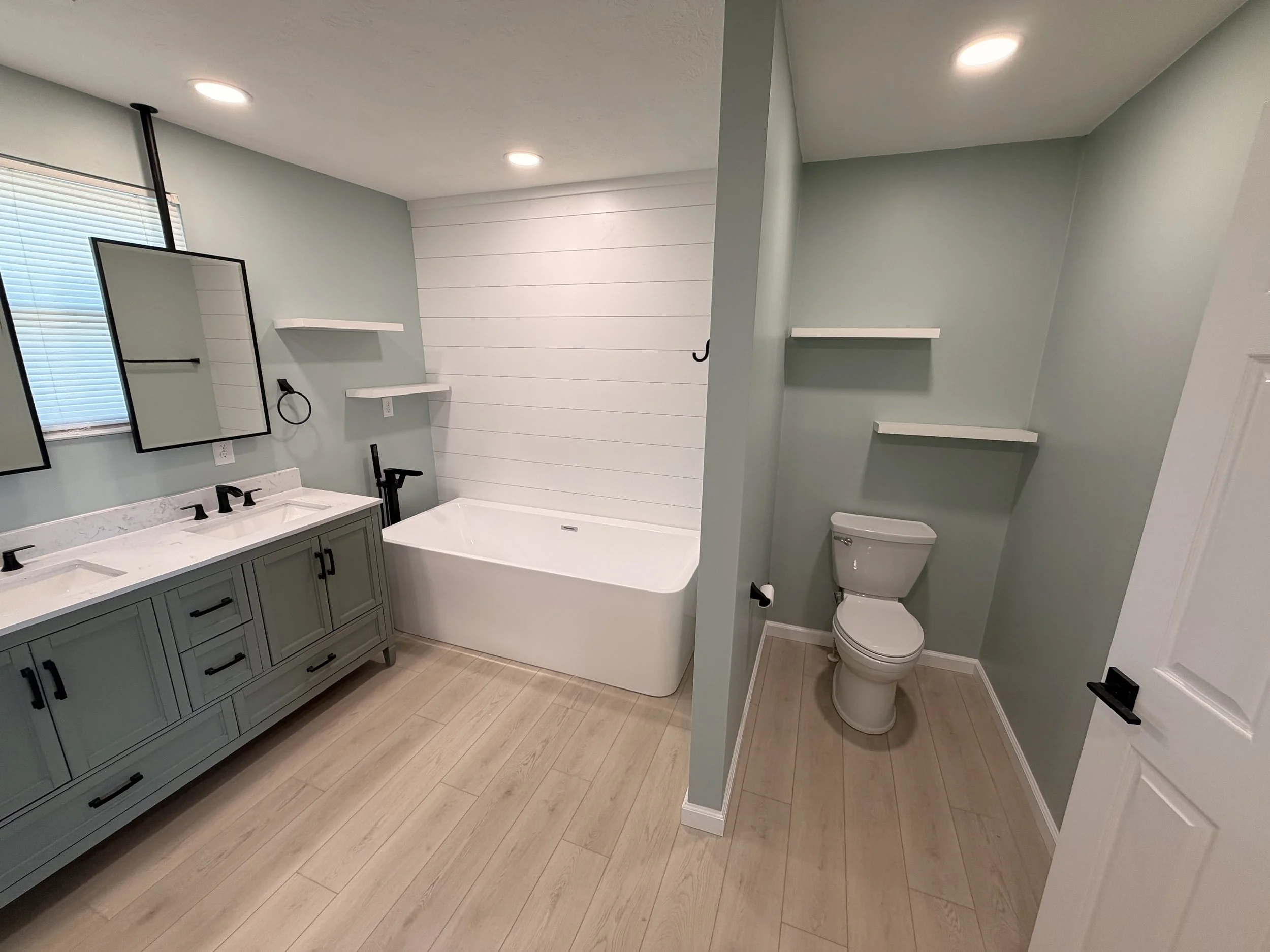 Modern bathroom with a double vanity with gray cabinets and black hardware, two sinks, and two mirrors. There is a bathtub with a white wood-paneled wall behind it. To the right, there is a toilet with a small wall shelf above it, and two small white